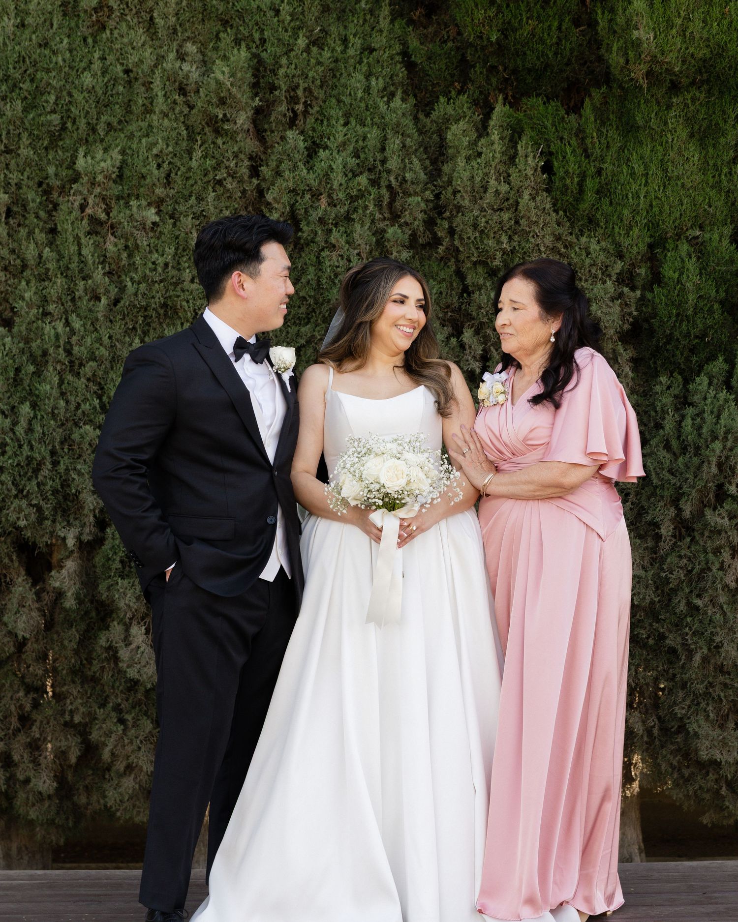 Bride in white gown holds bouquet flanked by groom in tuxedo and woman in pink dress before lush green hedge.