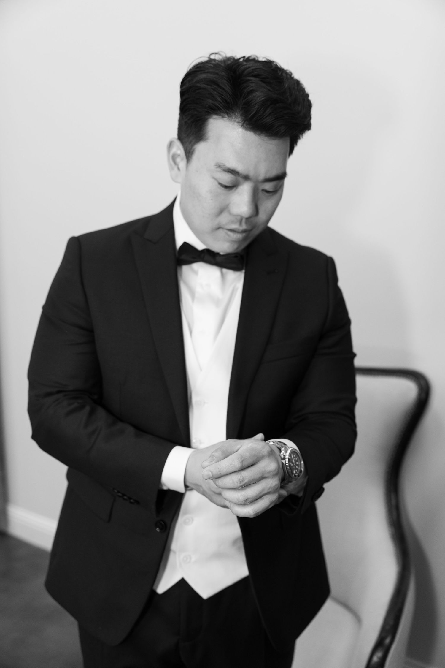 Groom in black tuxedo and bow tie adjusting his watch in a black and white photo.