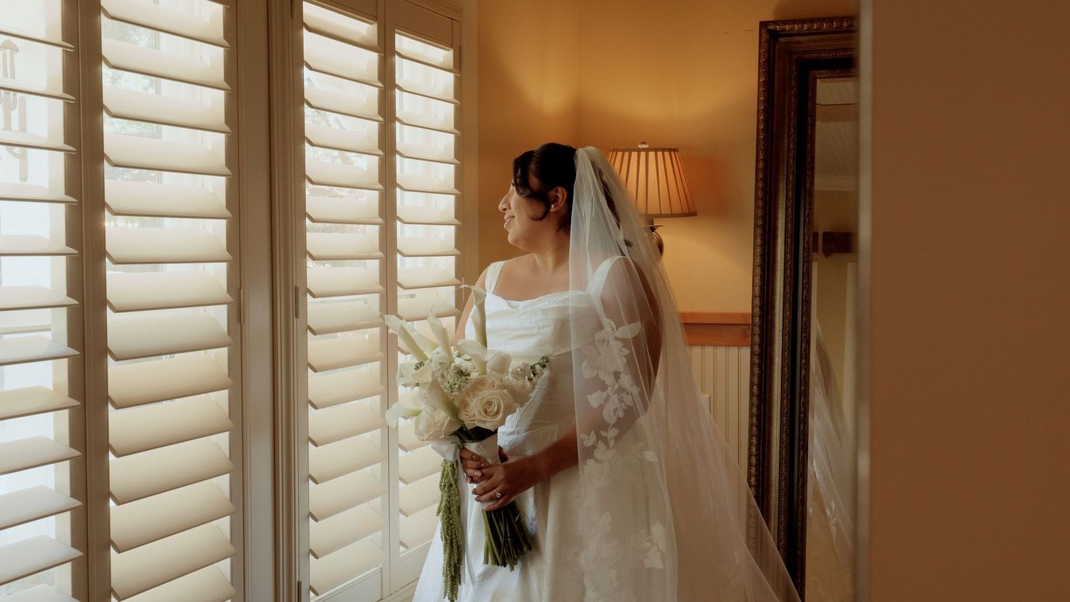A bride in a white lace gown and veil holds a bouquet near shuttered windows in a warmly lit room.