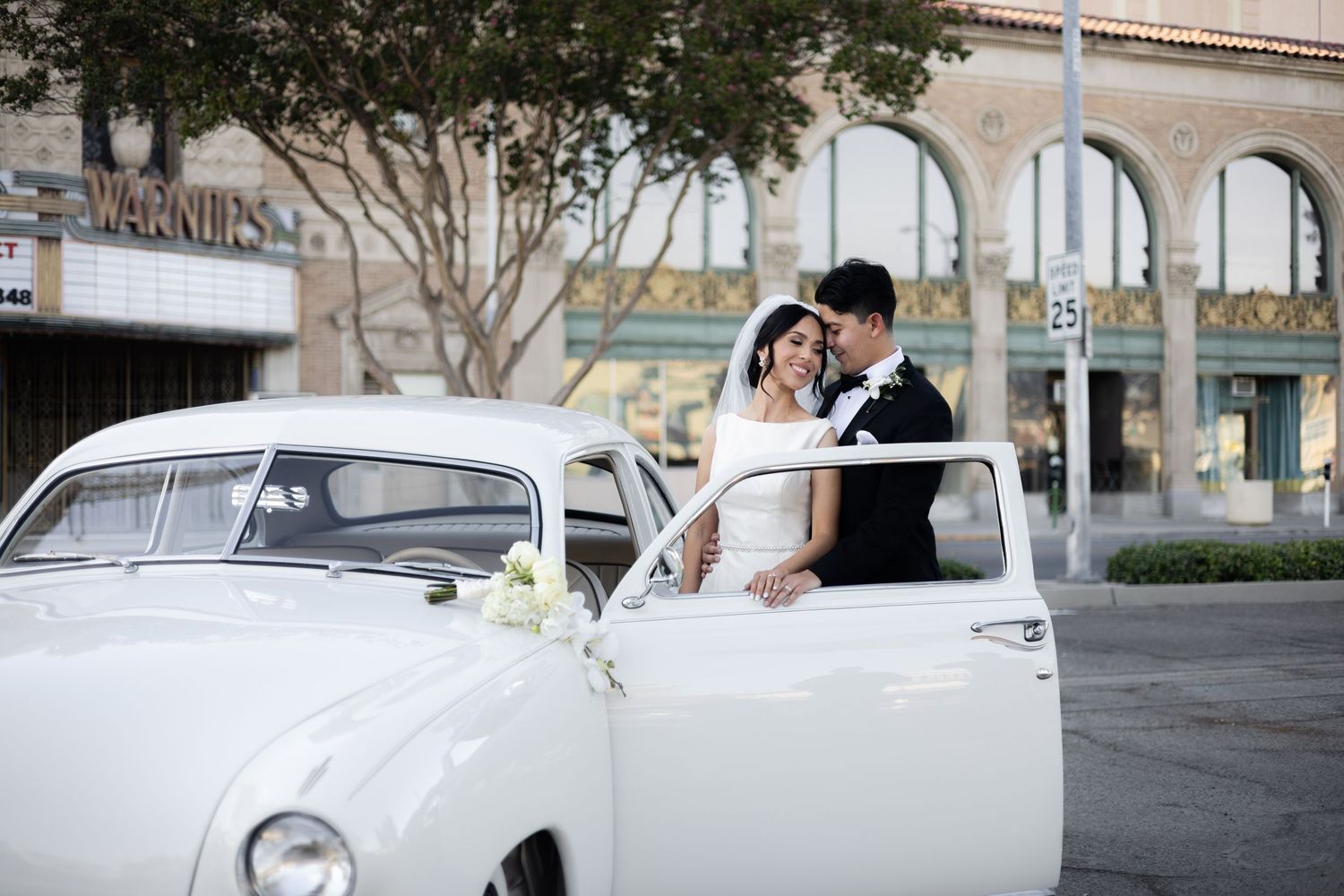 Vintage white car with wedding couple parked beneath trees on city street.