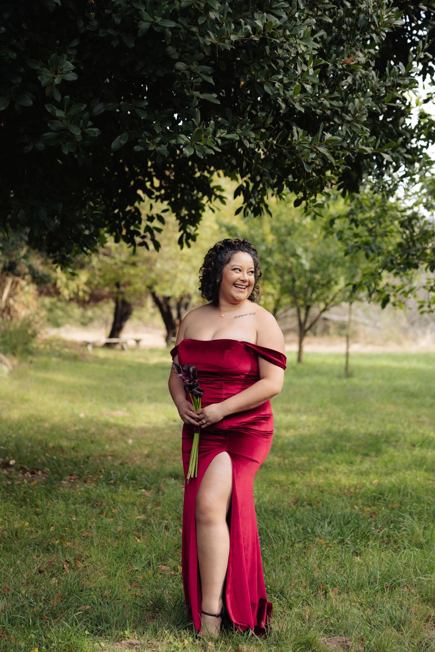 Individual in deep red formal dress poses confidently in a garden setting.