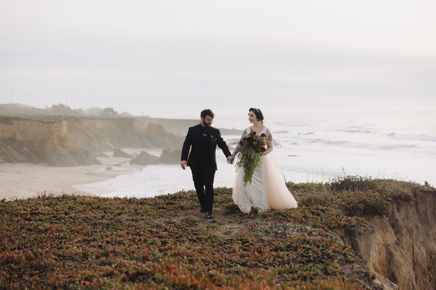 Newlyweds walk along dramatic coastal cliffs with crashing waves and overcast skies in the background.