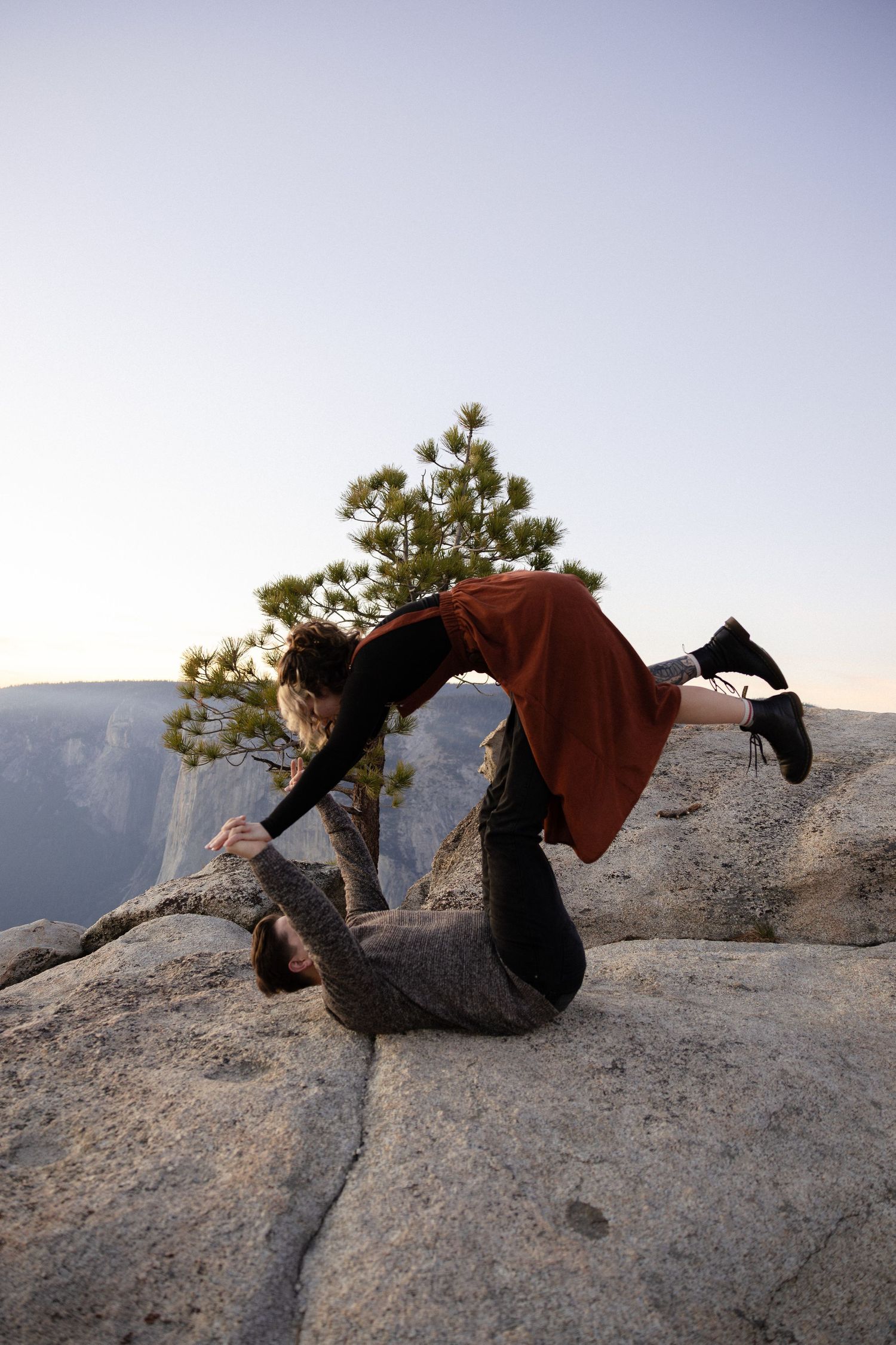 A couple lounges together on a rocky cliff edge overlooking a scenic mountain landscape at sunset.