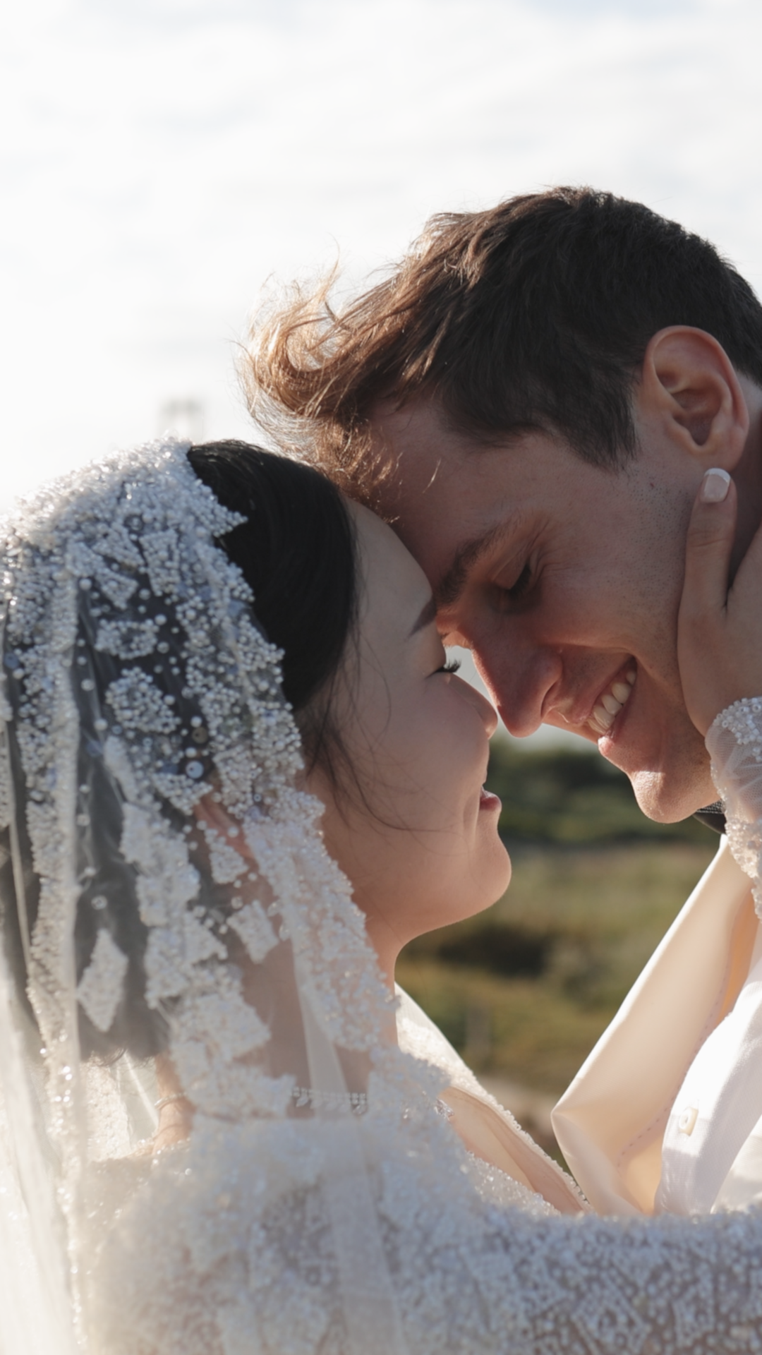 A smiling bride in a beaded veil and groom share an intimate forehead-to-forehead moment outdoors on their wedding day.