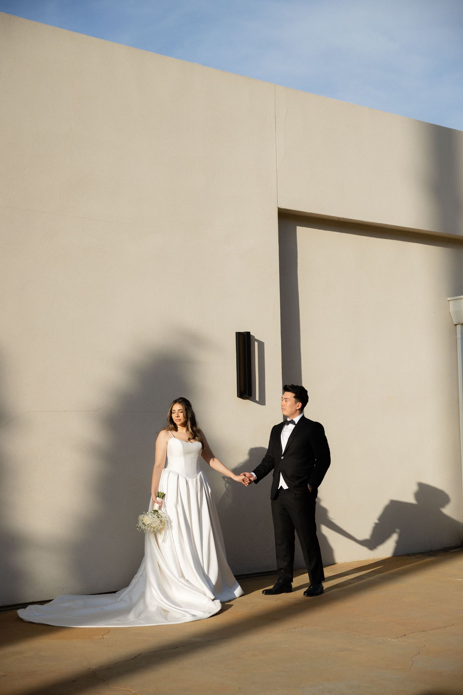 Bride in white gown and groom in tuxedo hold hands against a sunlit beige wall outdoors.