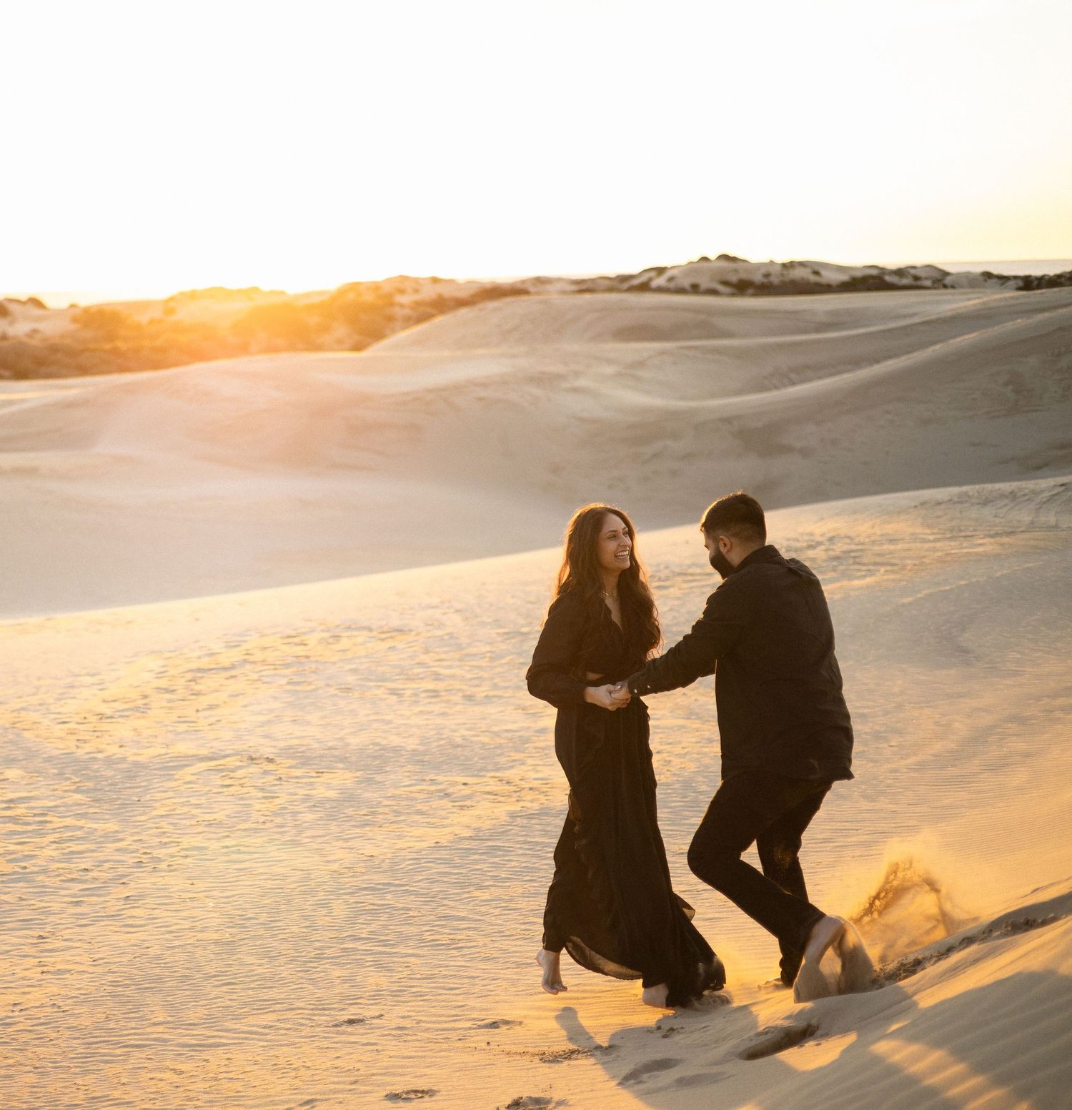 Couple sitting together on sandy dunes at sunset creating silhouettes against the golden sky.