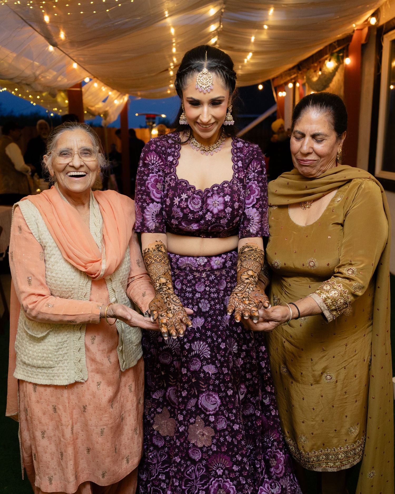 Three women in traditional Indian attire show their henna-decorated hands at a festive celebration under string lights.