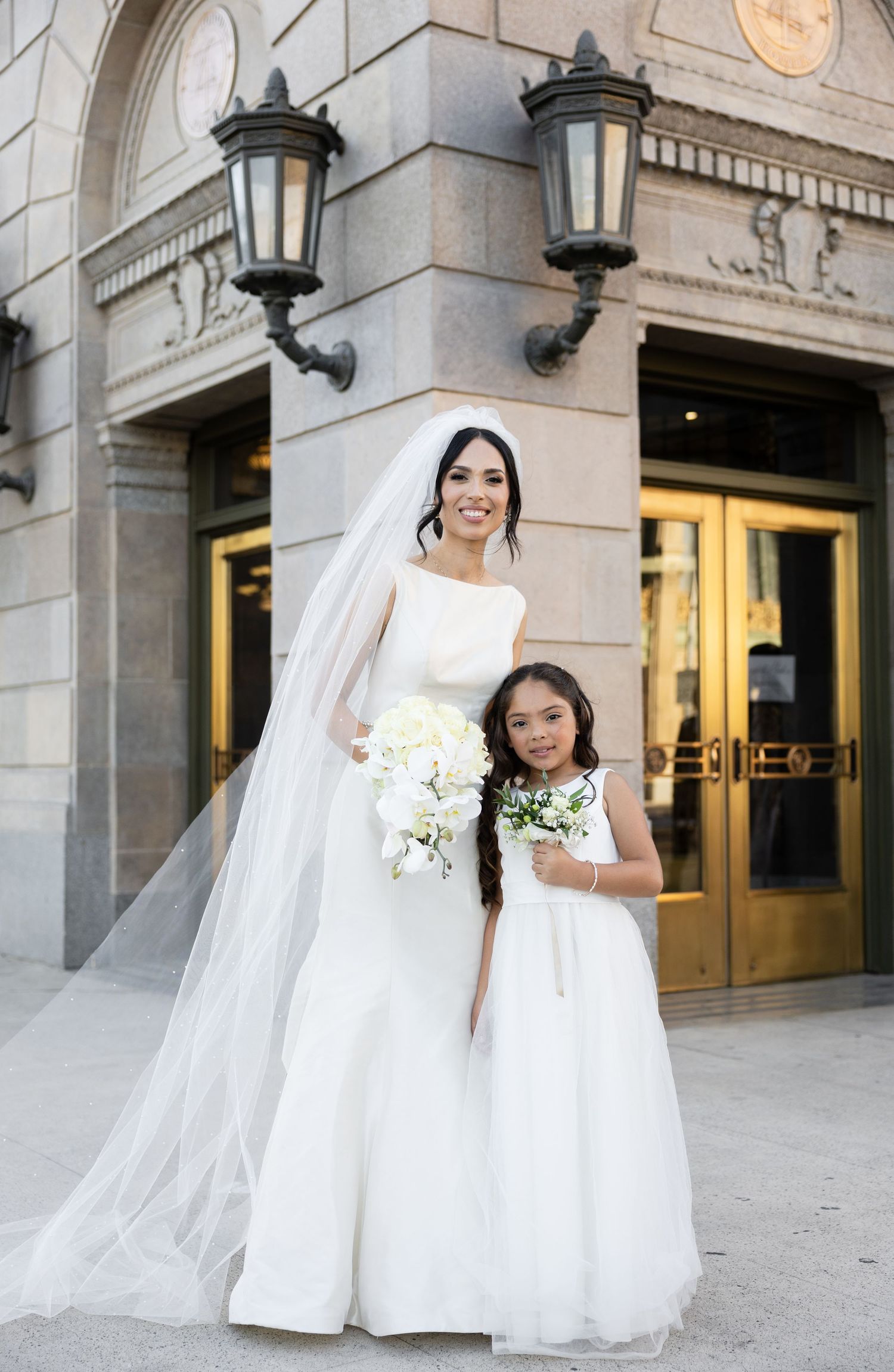 Elegant bride in white gown and flower girl in matching dress pose outside historic building with ornate lanterns.
