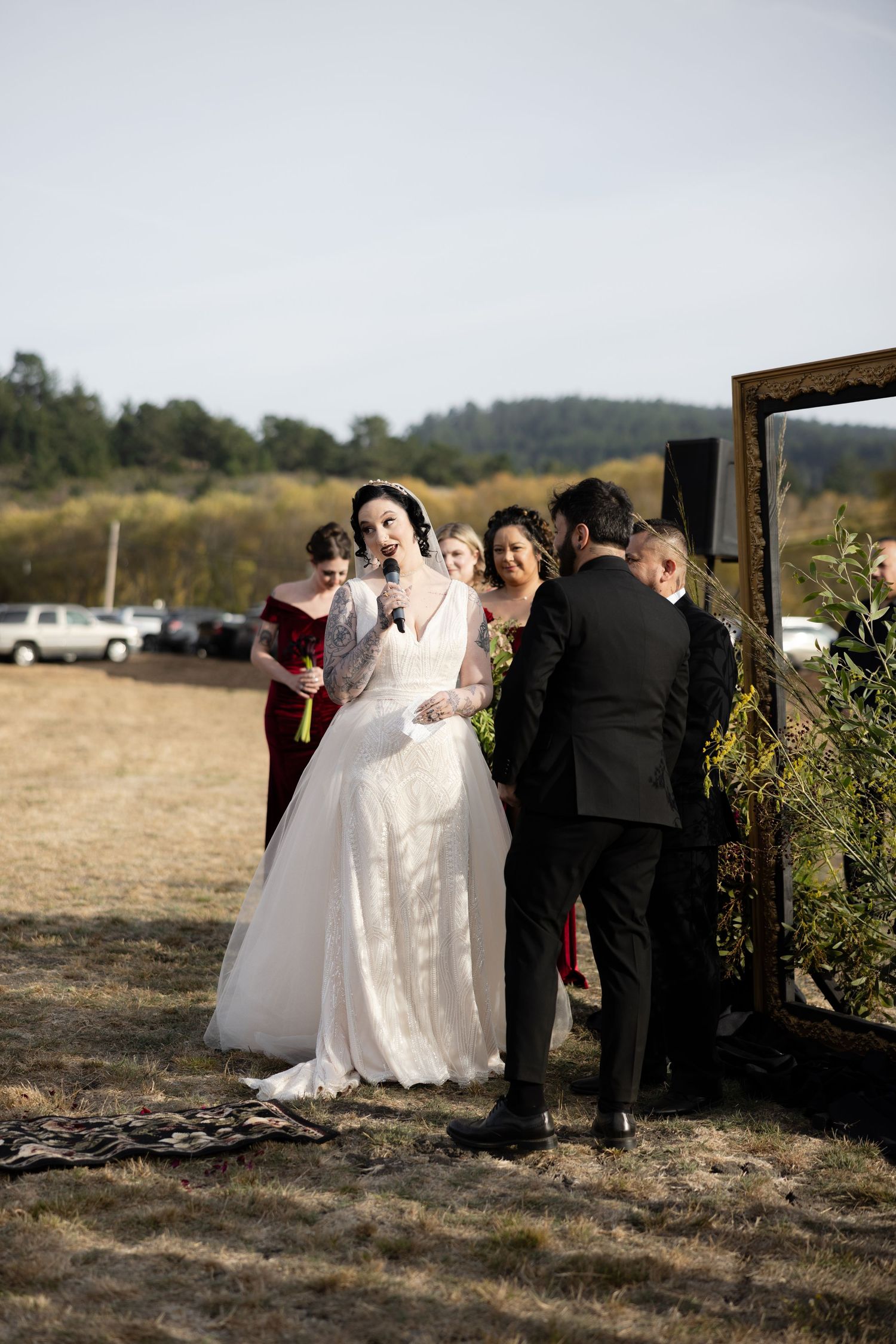 Wedding ceremony participants gather near wooden arch frame in rustic outdoor setting.