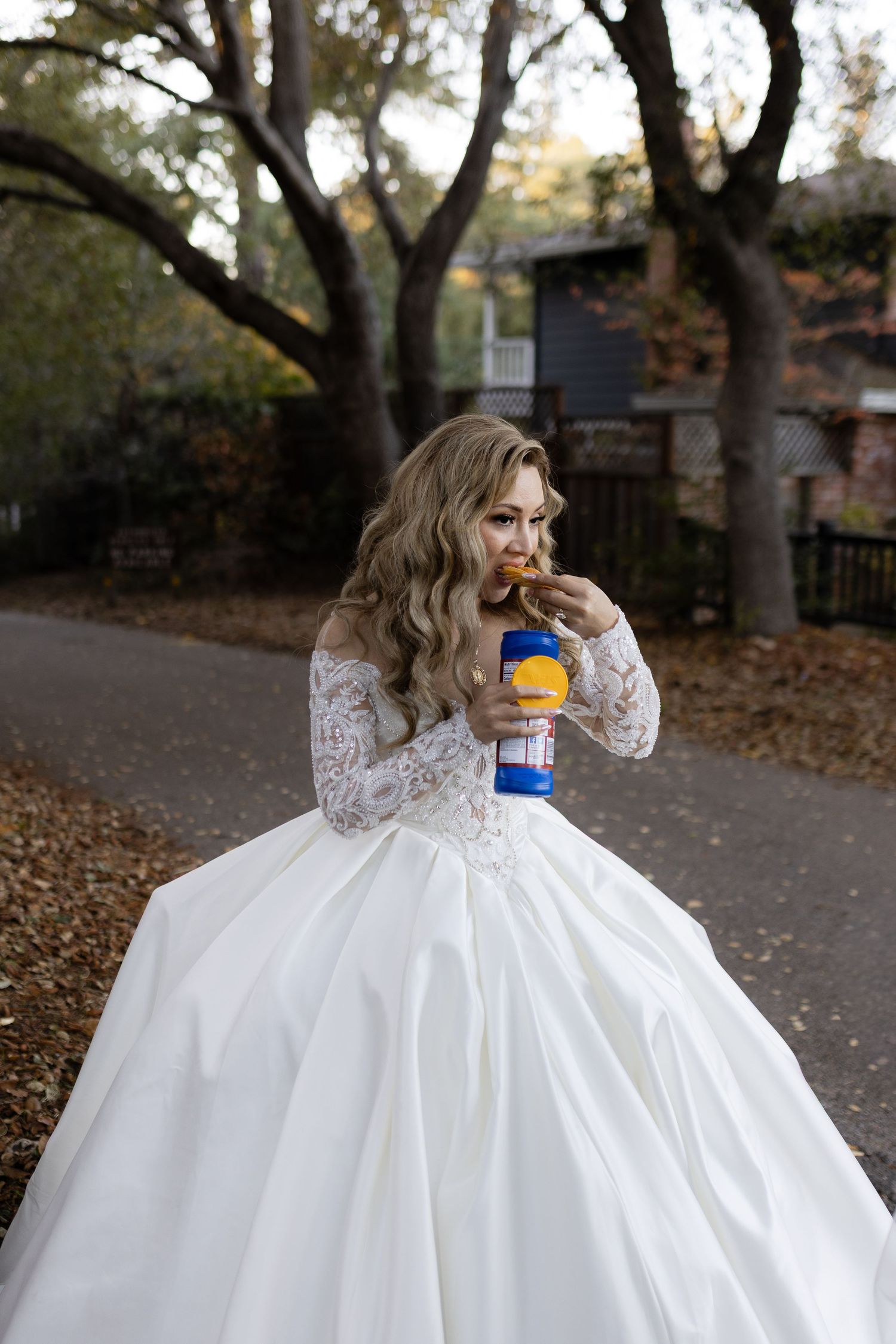 A bride in a voluminous white gown enjoys a drink outdoors among autumn trees.