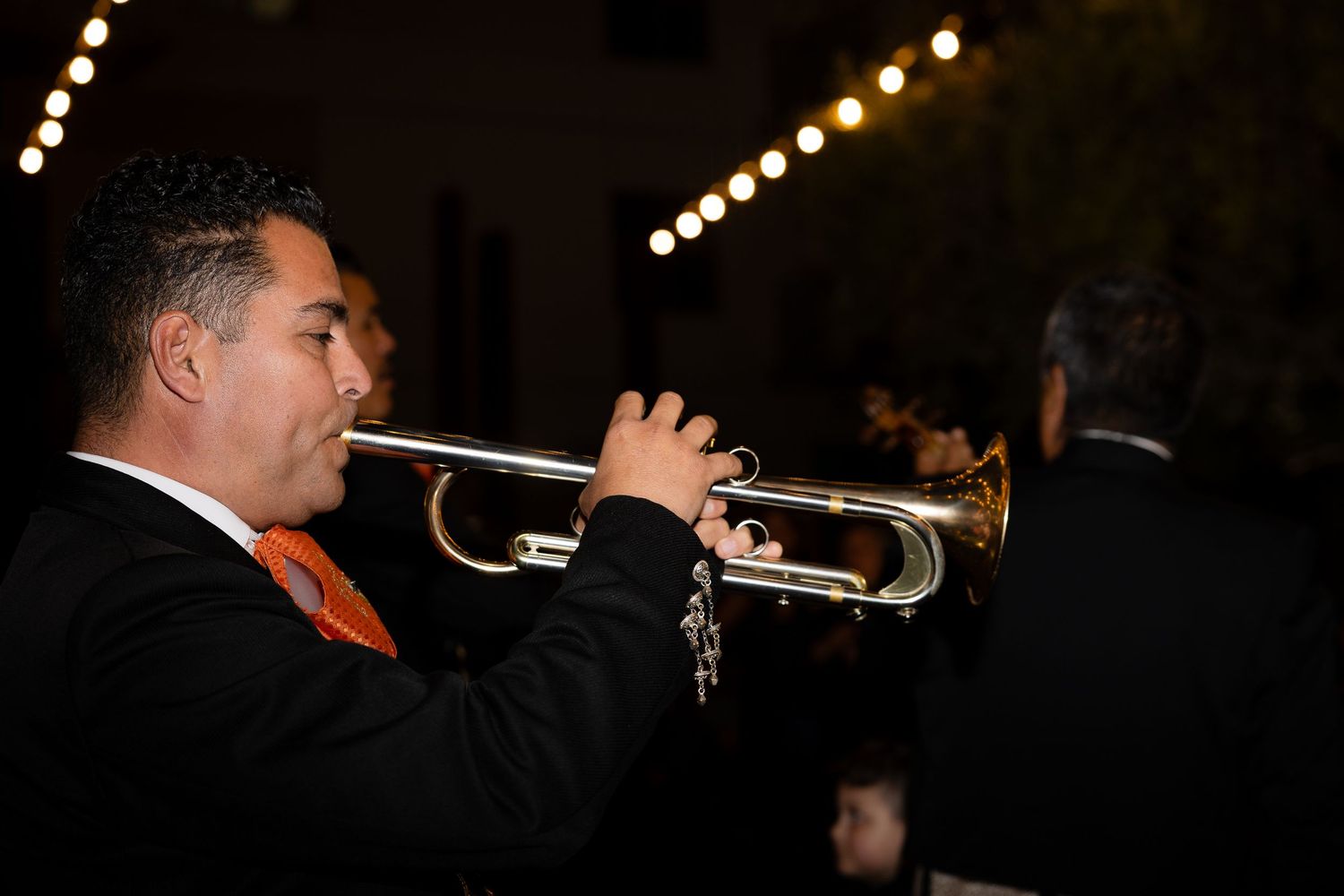 A trumpet player performs at a formal event in dark lighting with bokeh lights in the background.