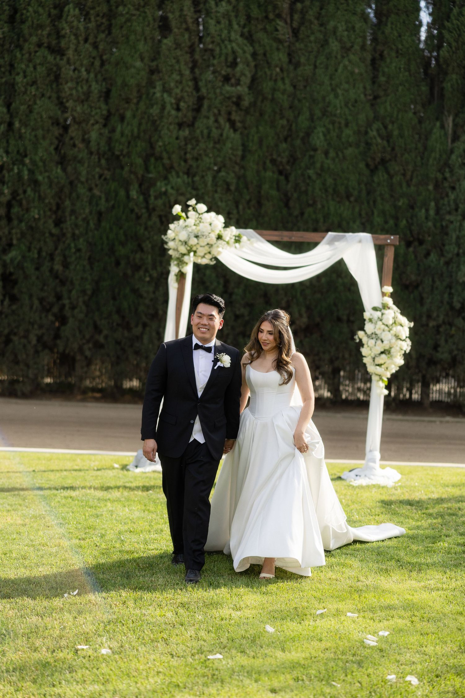 Smiling newlyweds walk hand in hand on green lawn in front of white floral wedding arch.