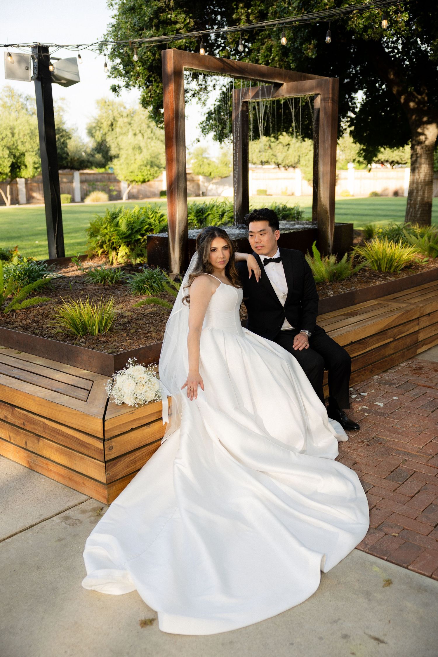 Bride in white ballgown and groom in black tuxedo pose together outdoors under a wooden arch at their wedding.