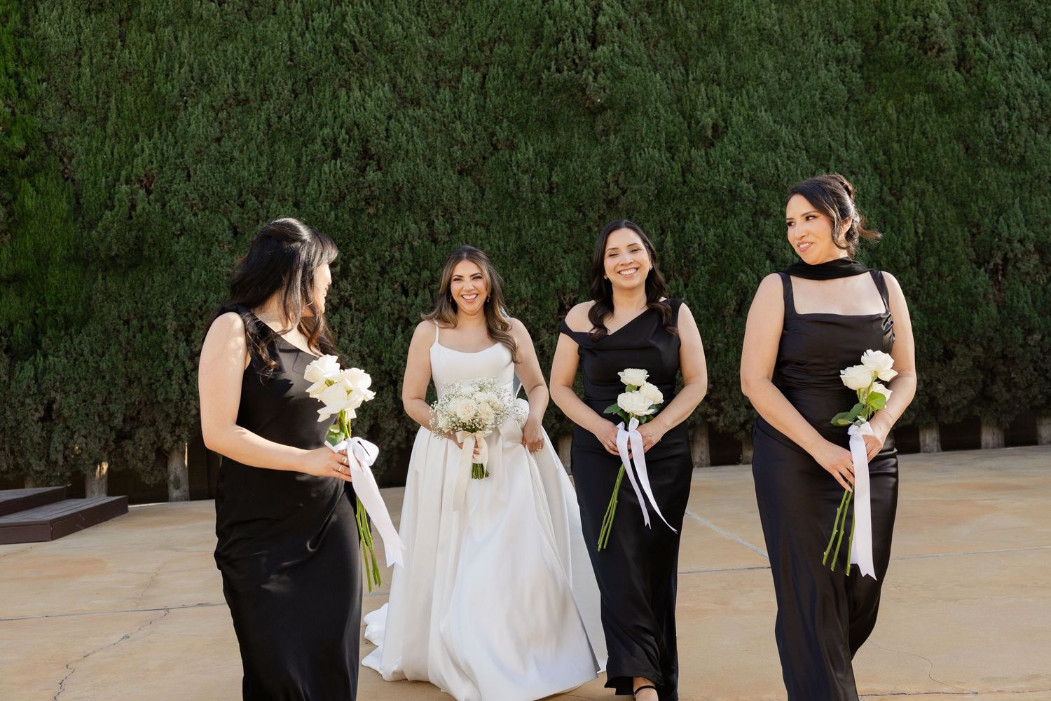 Bride in white gown with three bridesmaids in black dresses holding bouquets outdoors.