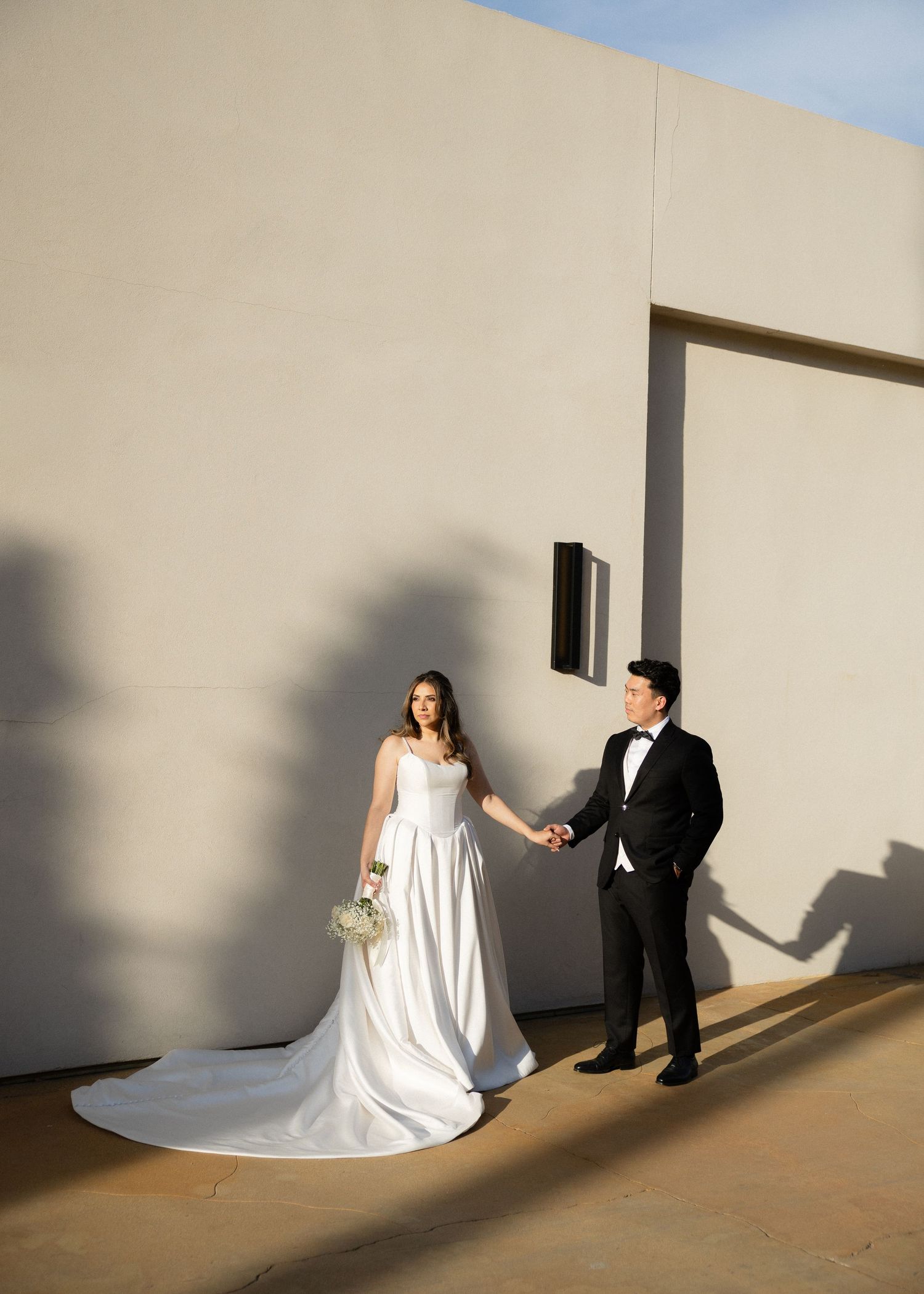 Bride in white gown and groom in tuxedo hold hands against a minimalist beige wall in elegant wedding portrait.