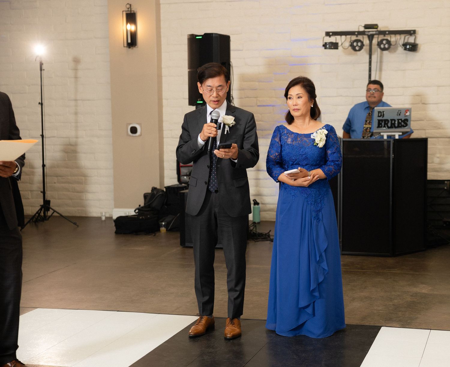Man in gray suit speaks into microphone beside woman in blue gown during wedding reception speech.