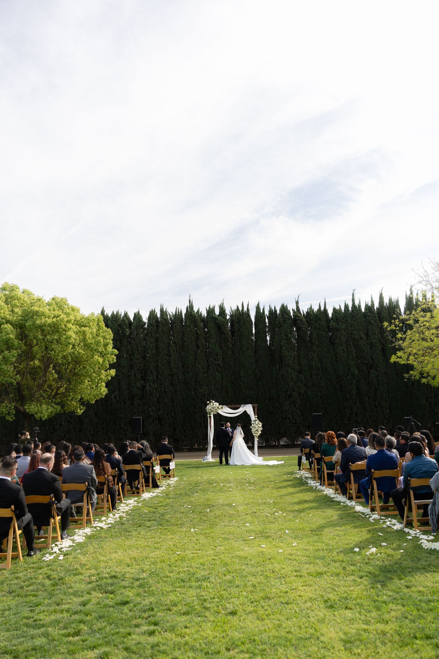 Wide shot of outdoor wedding ceremony on green lawn with guests seated and couple at floral arch.