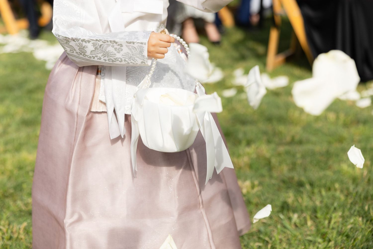 Flower girl in pink dress scatters white petals from basket during outdoor wedding ceremony.