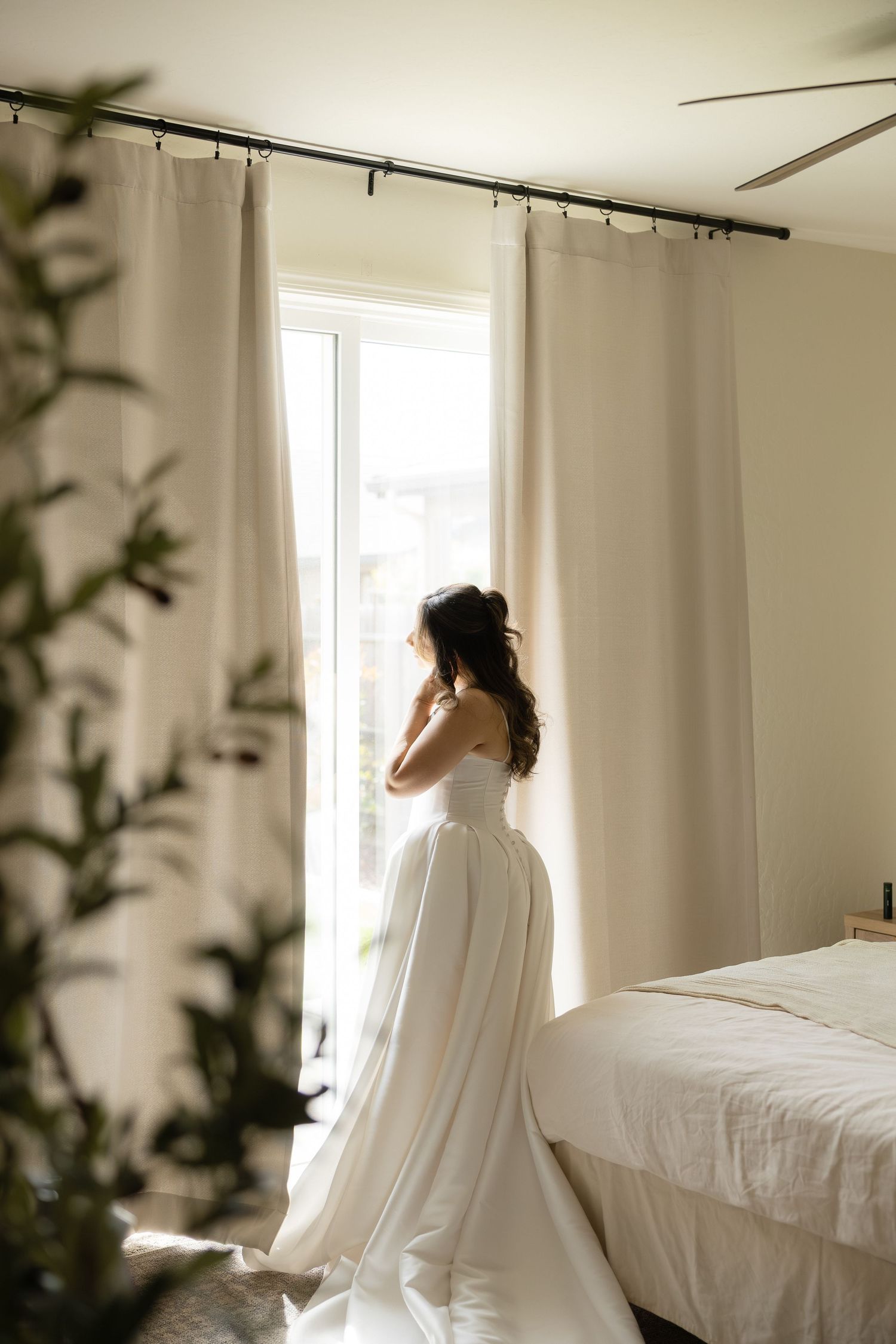 Bride in a white wedding gown standing by a sunlit window in a bright bedroom before her ceremony.