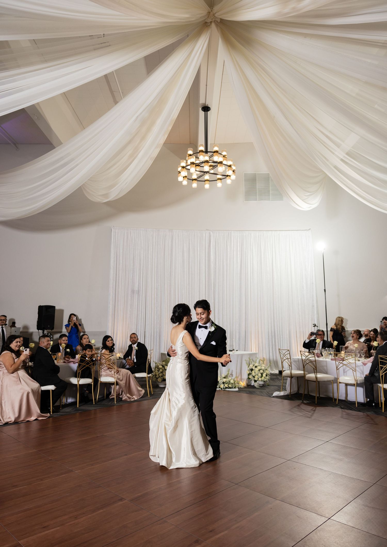 Wedding reception ballroom with dramatic white ceiling draping and guests watching first dance.