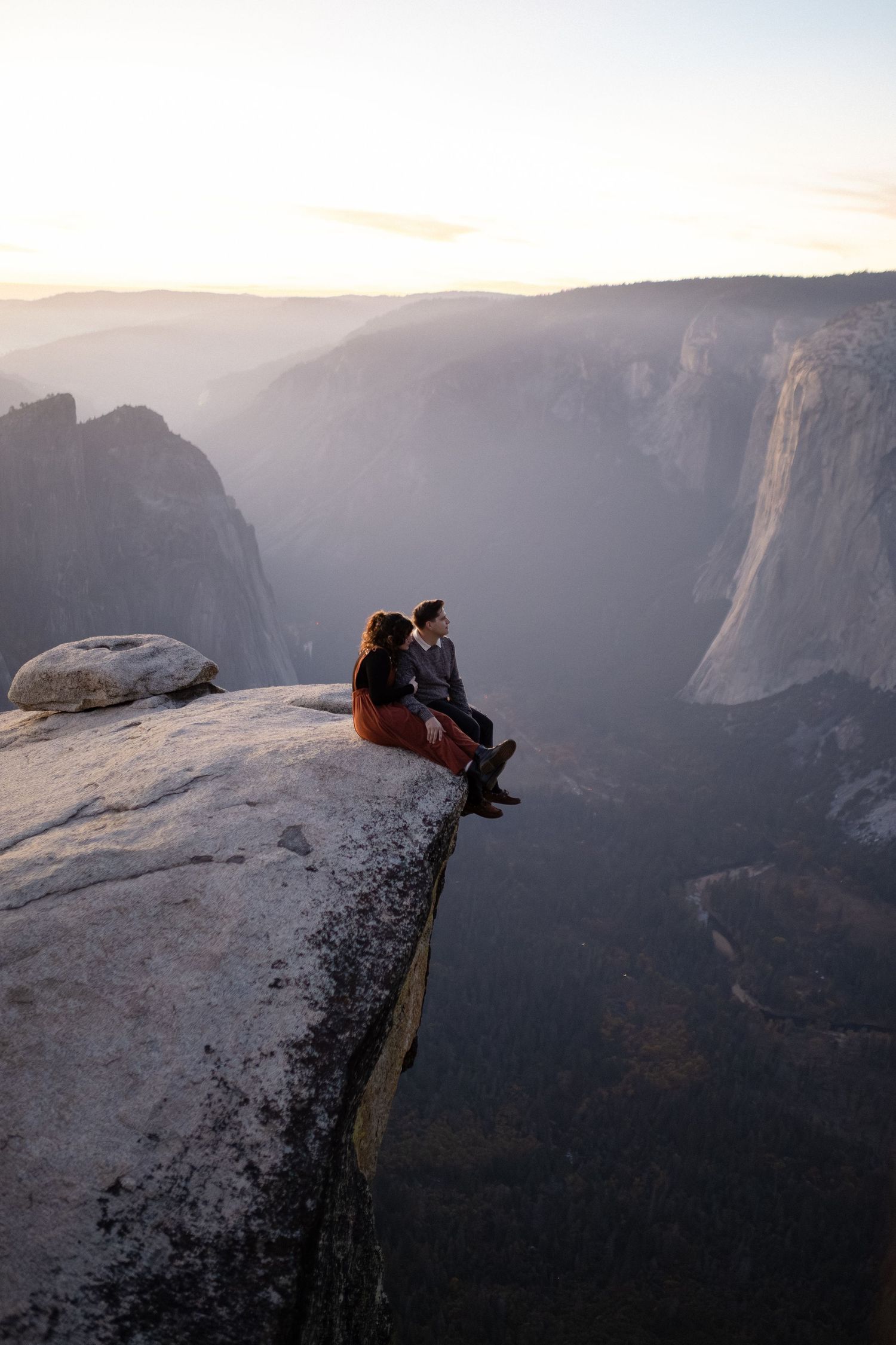 Silhouettes of people sitting on cliff edge against misty mountain backdrop in evening light.