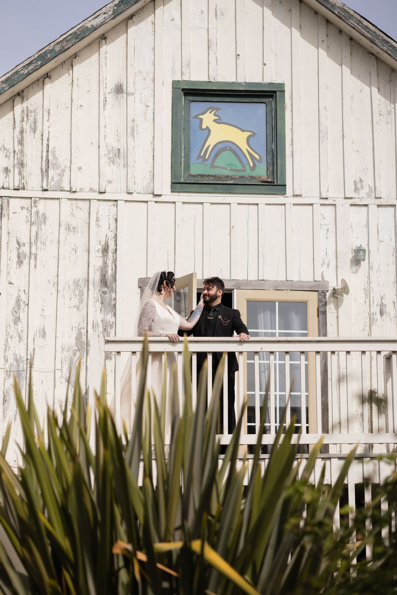 A couple converses on a white barn's balcony with decorative deer artwork and desert plants below.