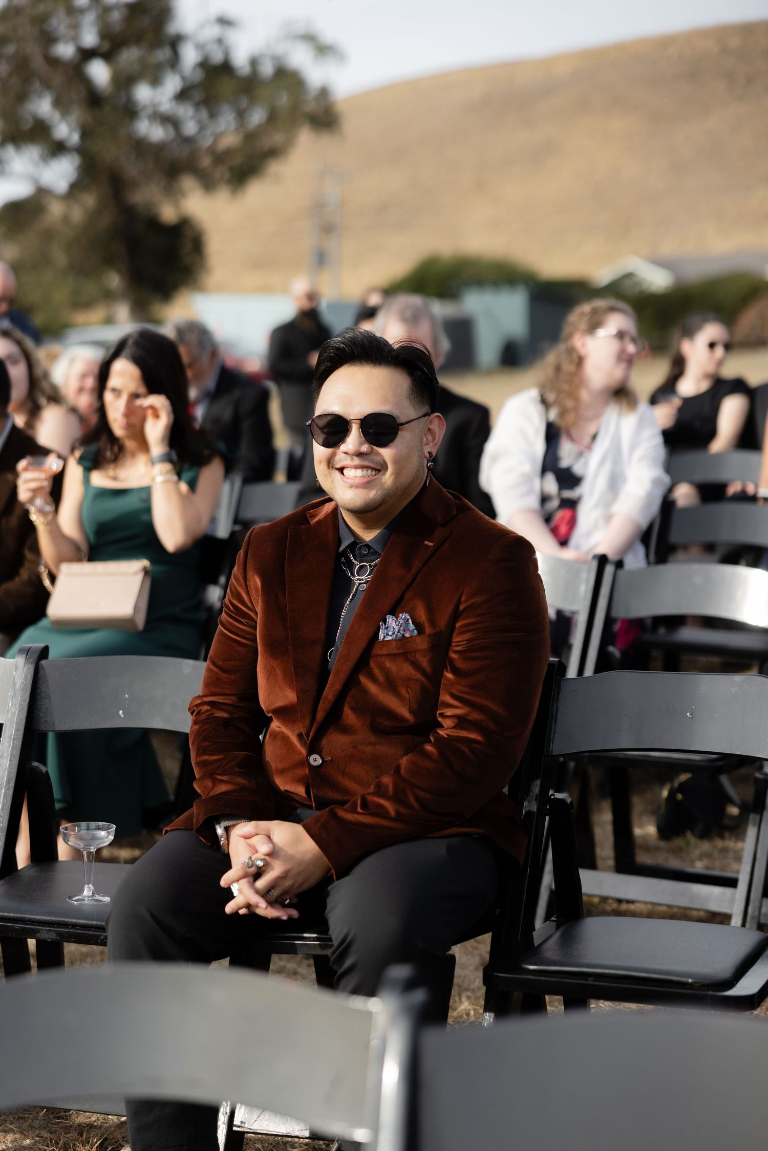 Wedding guests seated in black chairs at outdoor ceremony venue with rolling hills backdrop.