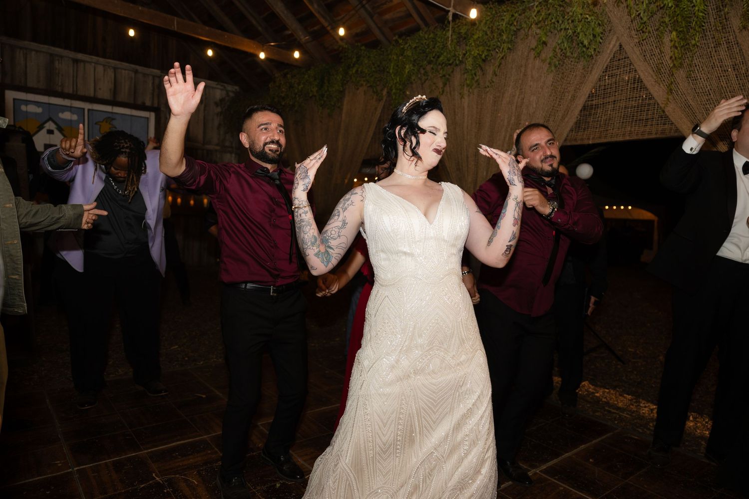 Wedding celebration with guests dancing and raising hands under decorative greenery and lights.
