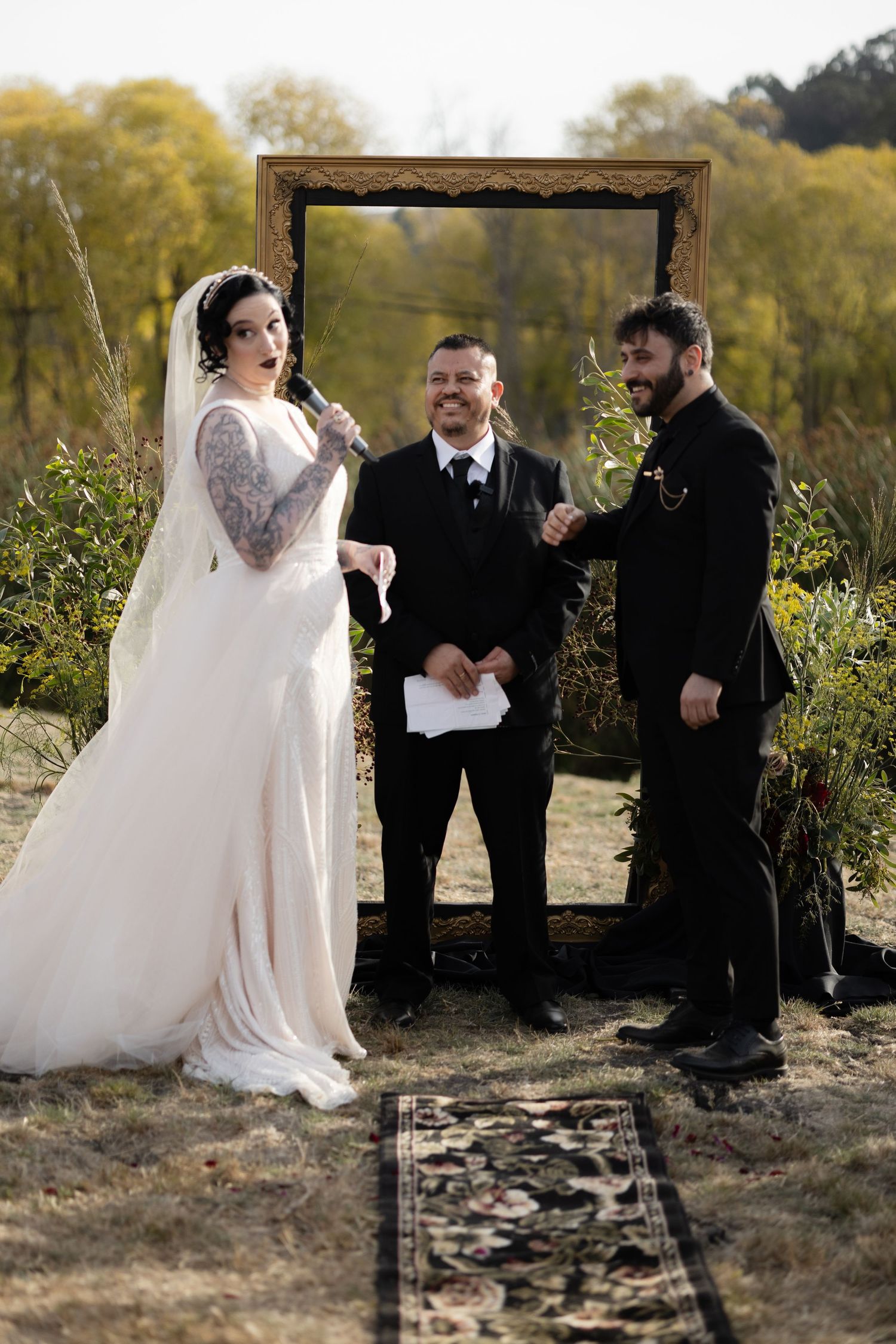 A wedding ceremony unfolds beneath a wooden frame arch in a natural outdoor setting.