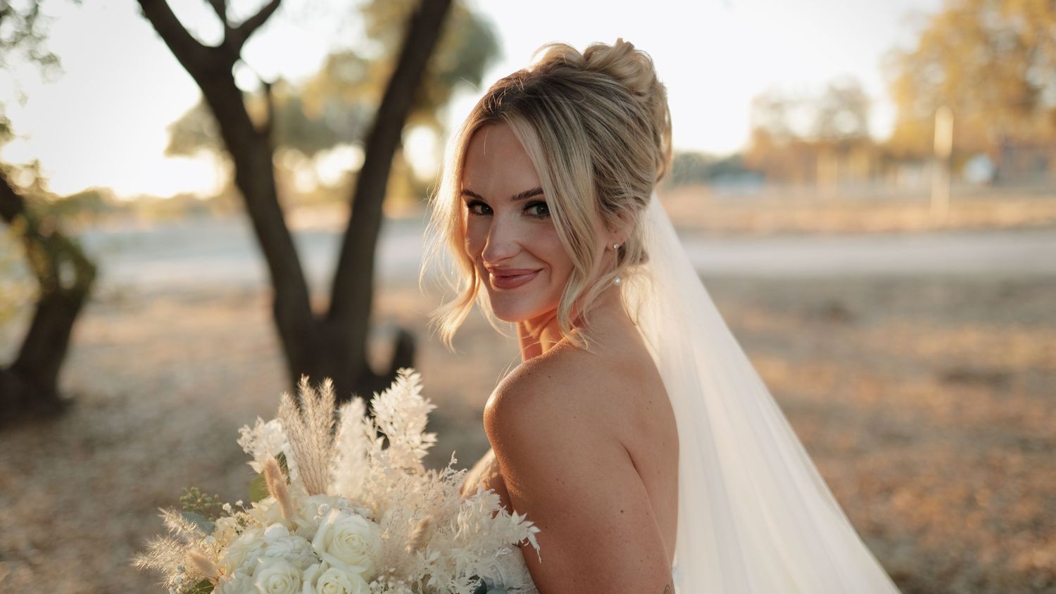 Smiling bride in white gown and veil holding a bouquet of white flowers outdoors at golden hour.