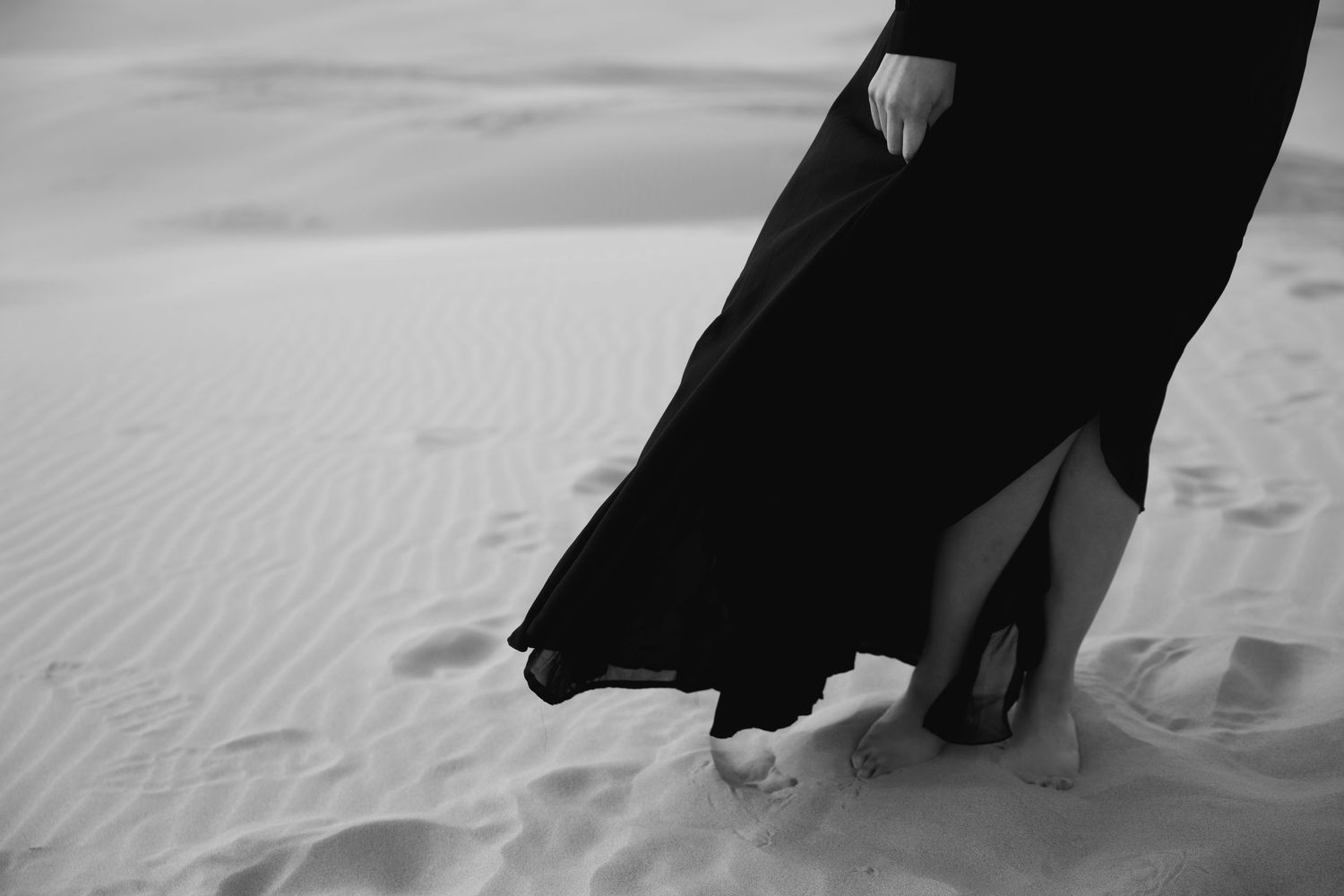 A couple in formal attire walks on a sandy beach, with their feet leaving footprints in the white sand.