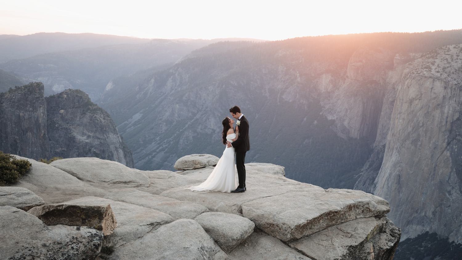 Bride and groom share a kiss on a rocky cliff edge at sunset in Yosemite National Park with valley views.