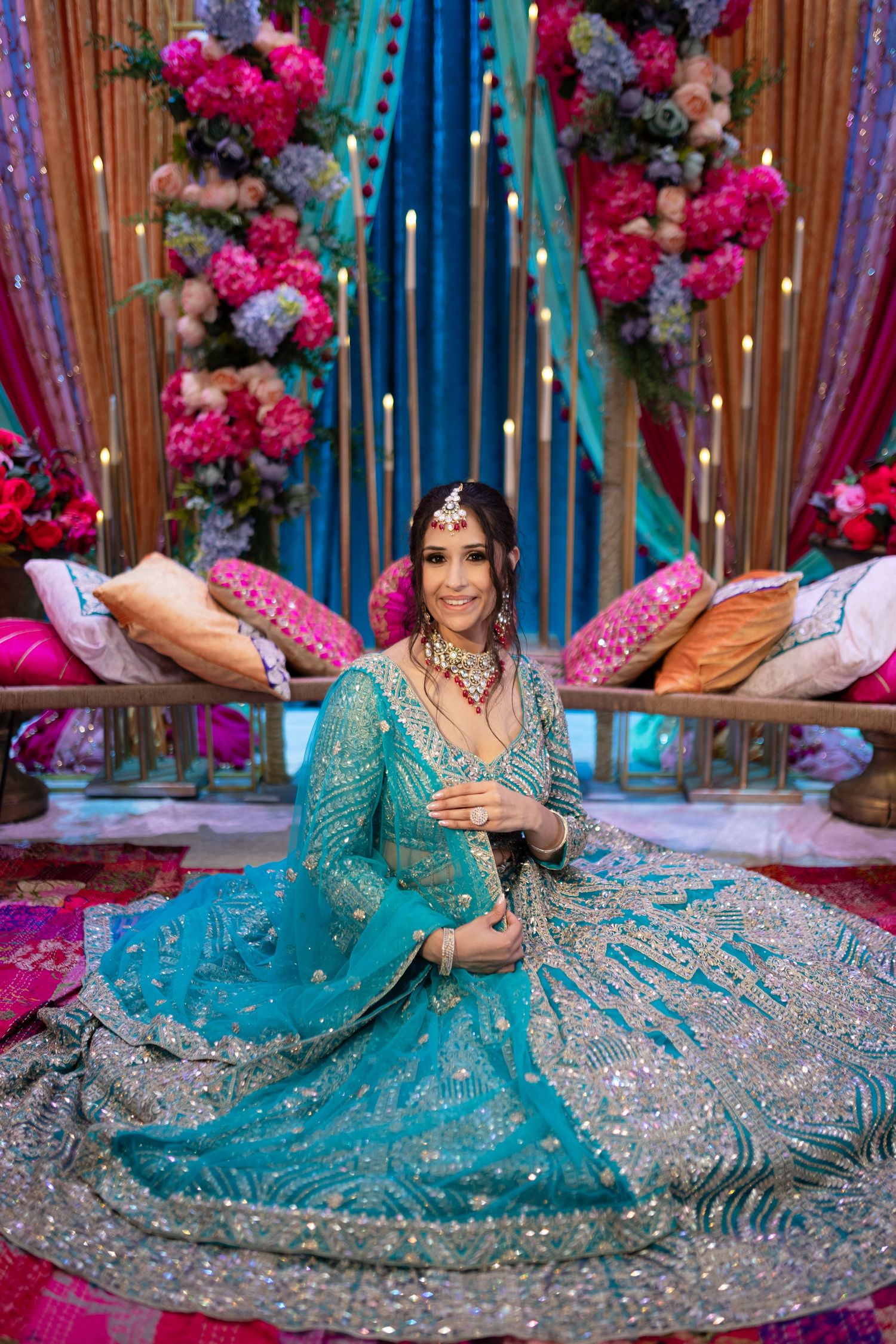 A person in a turquoise and silver embellished lehenga poses gracefully on the floor with floral decorations.
