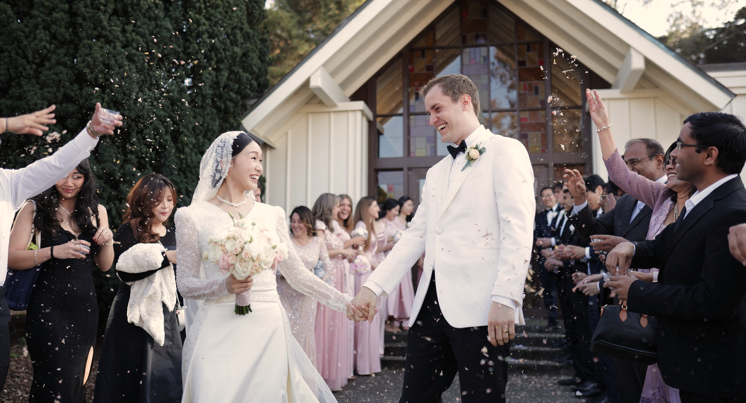 Bride and groom exit chapel amid confetti shower, surrounded by wedding party in pink dresses and black suits.