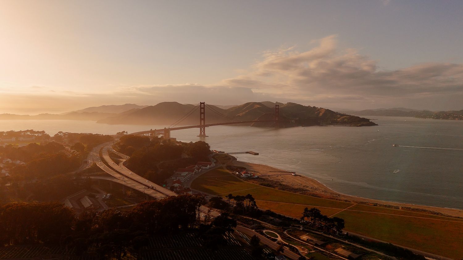 Aerial view of the Golden Gate Bridge at sunset with warm golden light over San Francisco Bay and rolling hills.