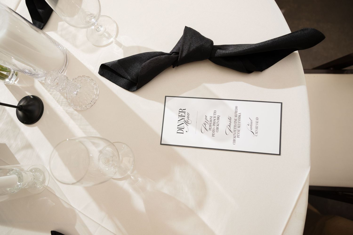 Elegant dinner table setting with black bow tie, menu card, and crystal glasses on white tablecloth.