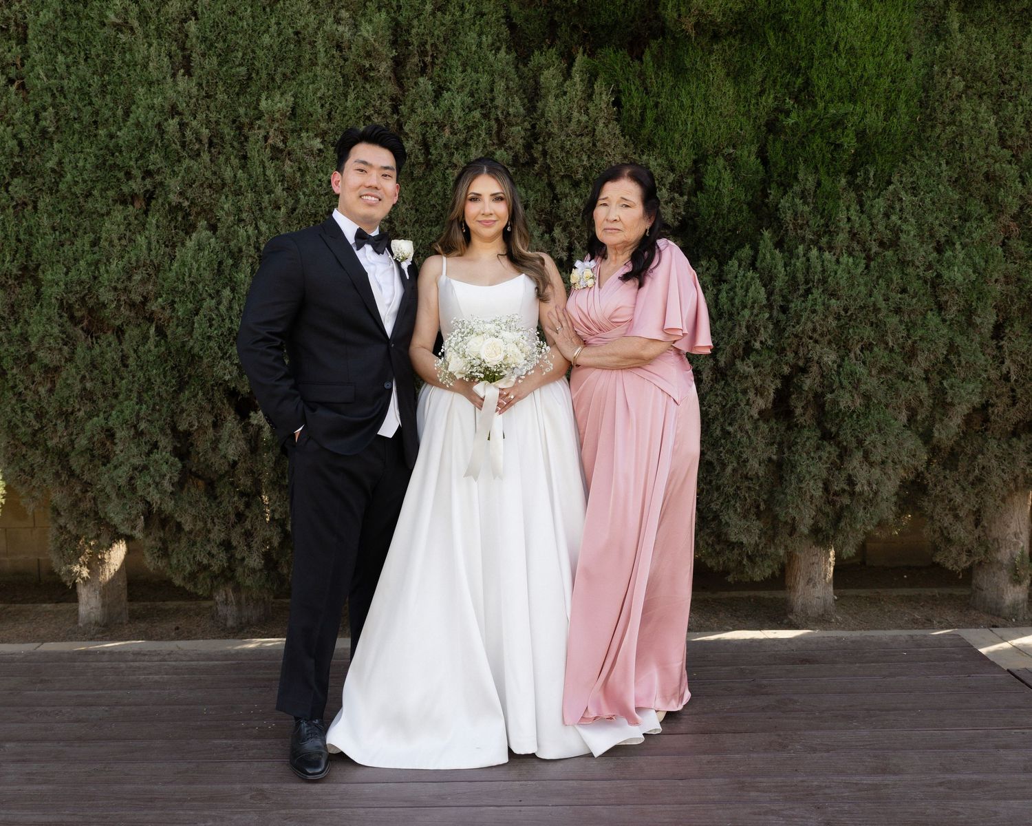 Bride in white gown poses with groom in tuxedo and woman in pink dress before lush green hedge backdrop.