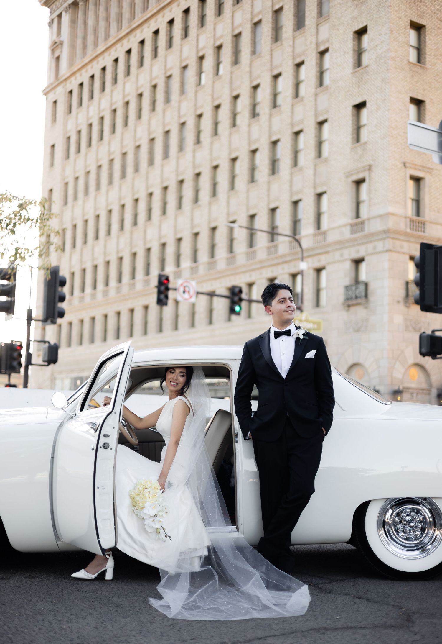 Classic white vintage car parked beside a grand stone building with wedding couple posing elegantly.