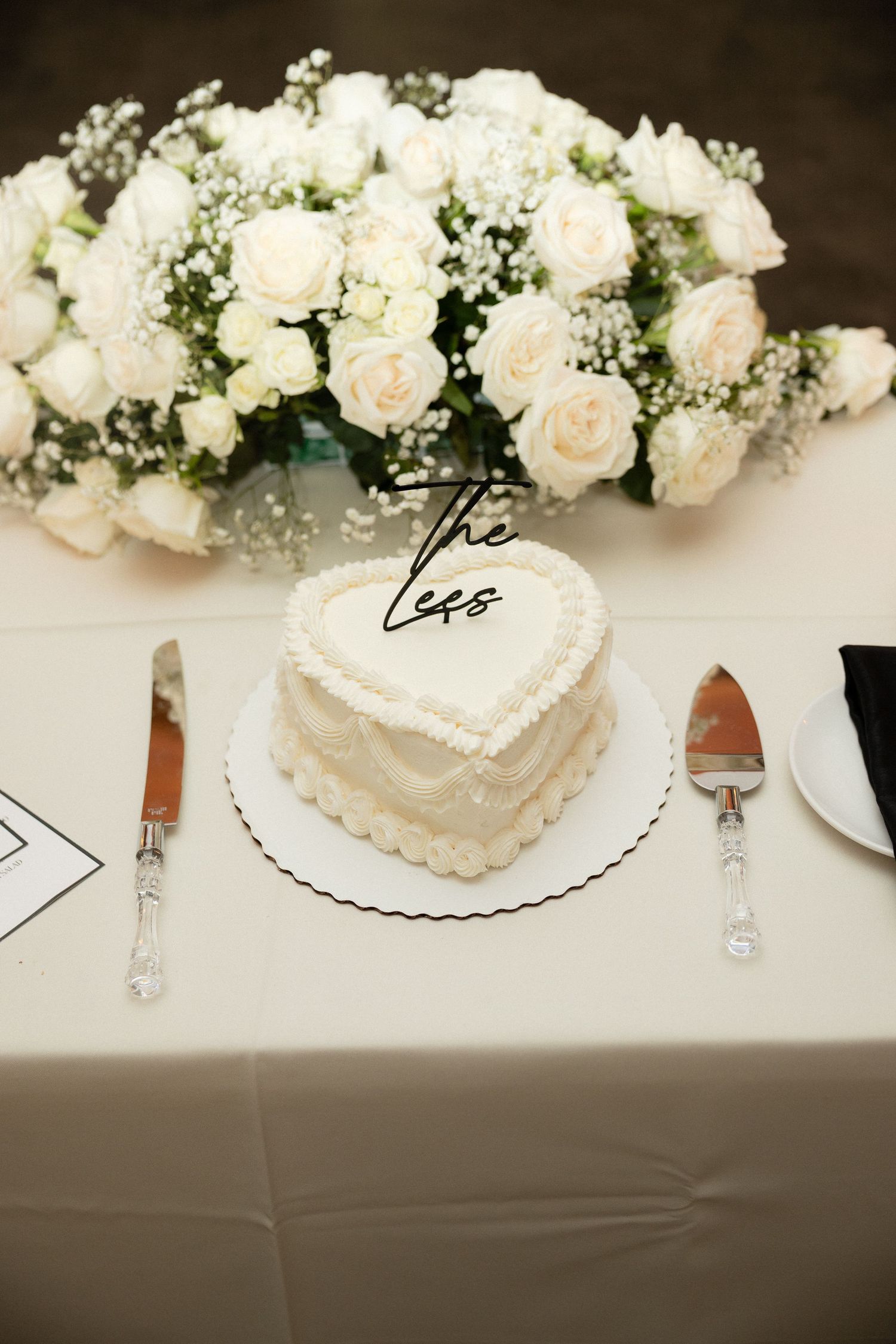 Heart-shaped white wedding cake displayed on table beside cutting knife and white rose floral arrangement.