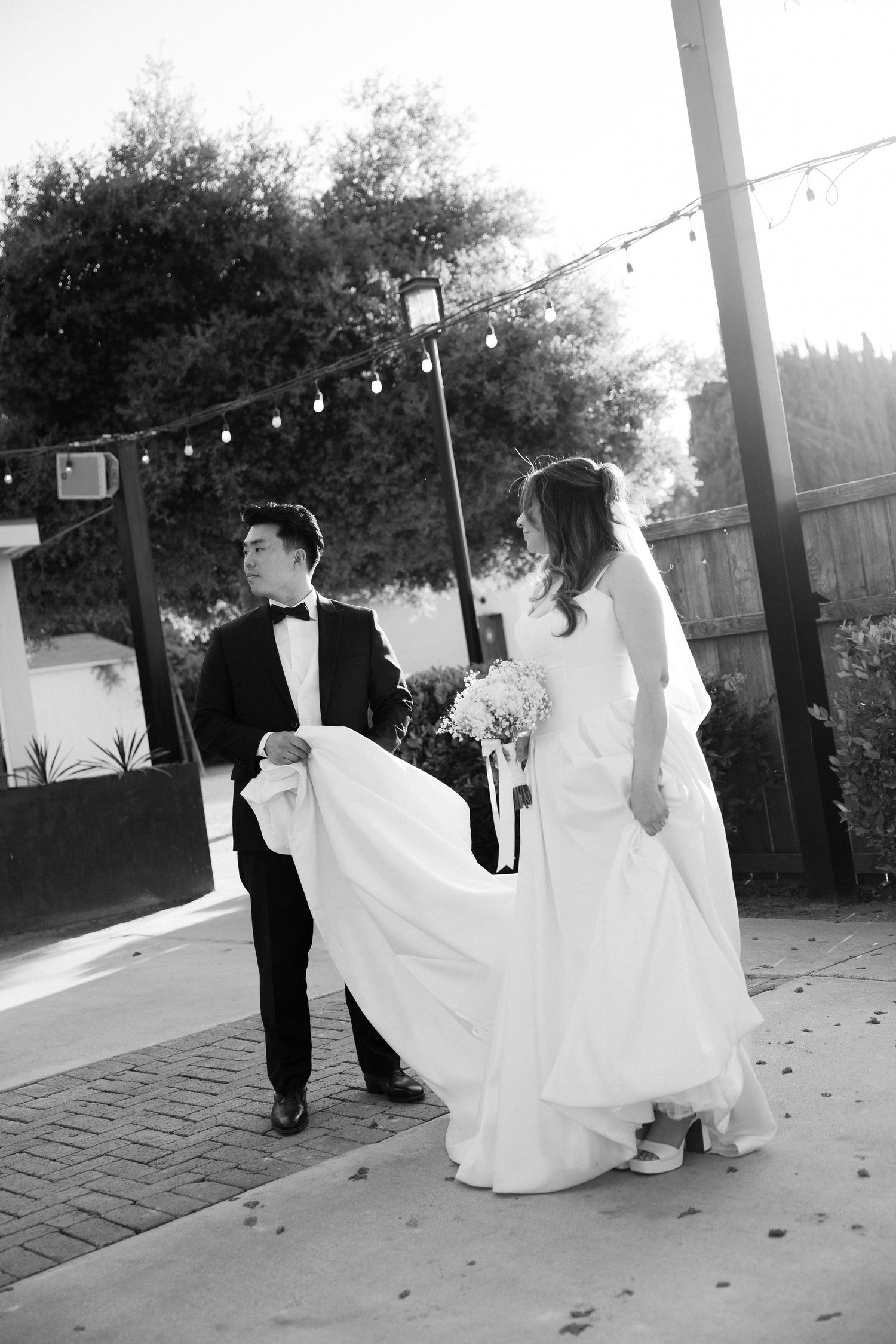 Black and white photo of a bride and groom walking together outdoors, groom holding the wedding dress train.