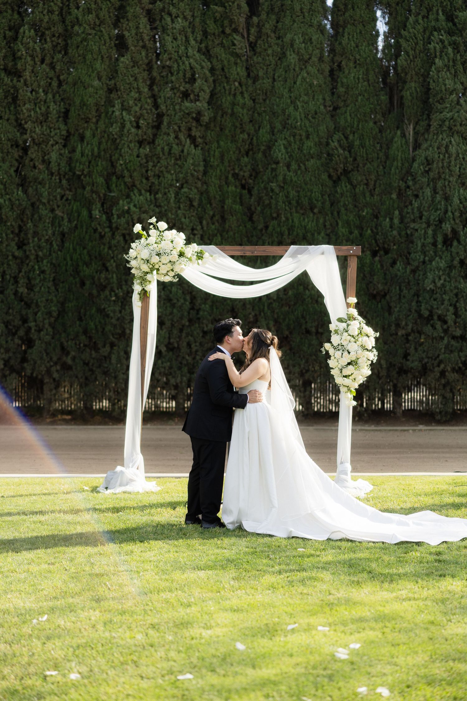 Bride and groom share first kiss at decorated outdoor wedding arch on green lawn.