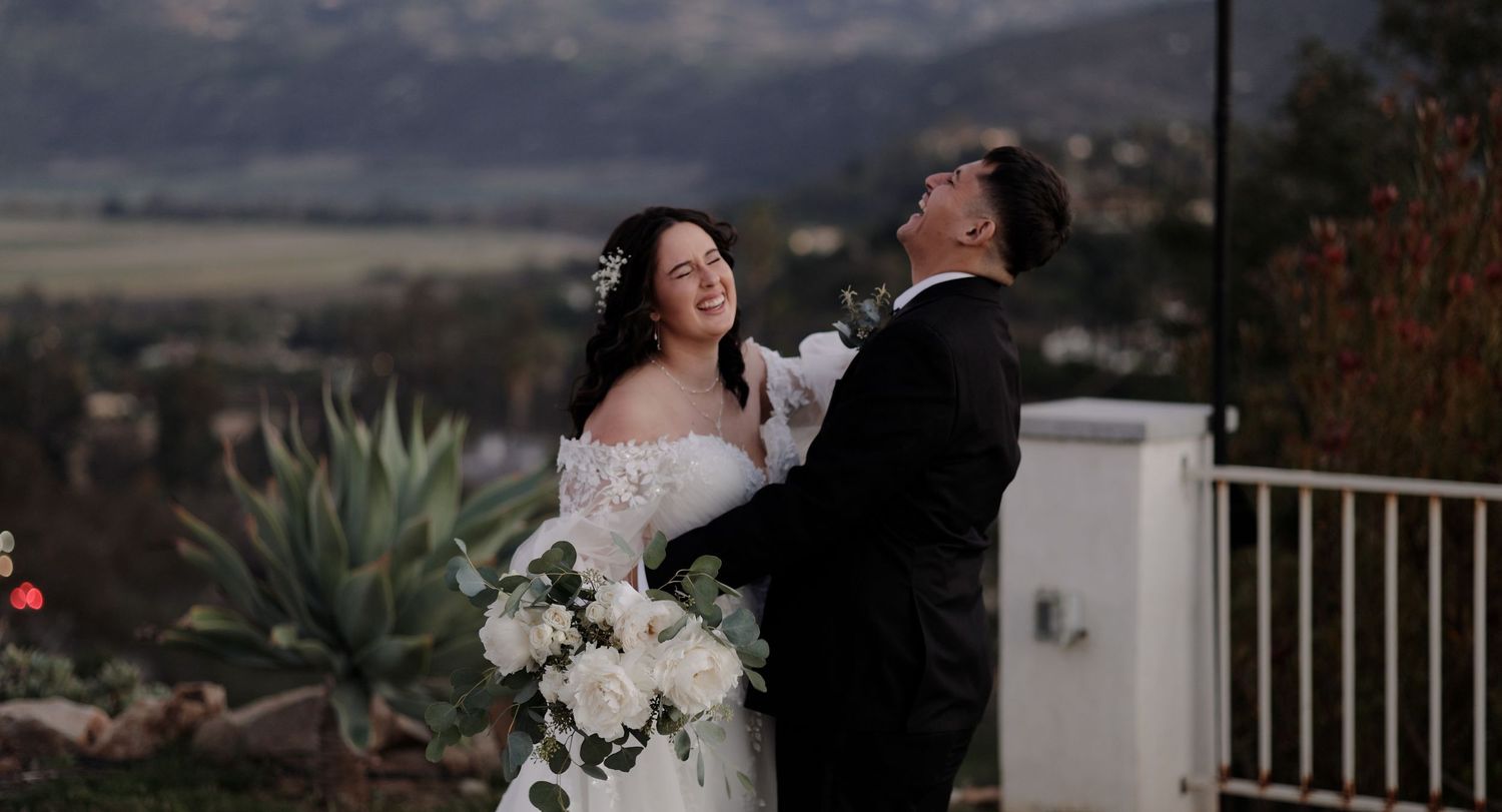 Bride and groom share a joyful moment outdoors with mountain views, she holds white floral bouquet in wedding attire.