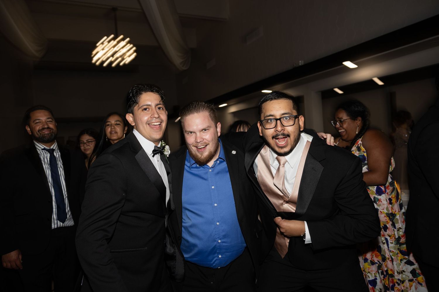 Three guests posing together at a wedding reception with decorative lighting overhead.