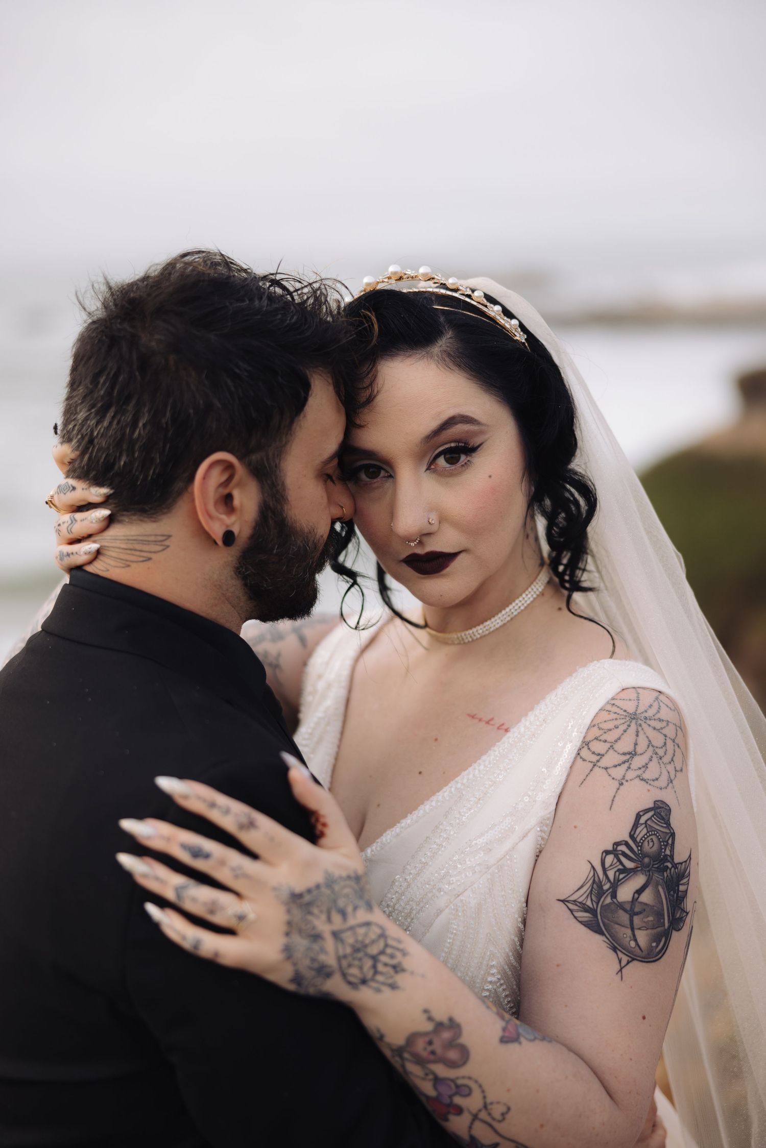Gothic-inspired wedding portrait featuring dramatic makeup and tattoos against coastal backdrop.