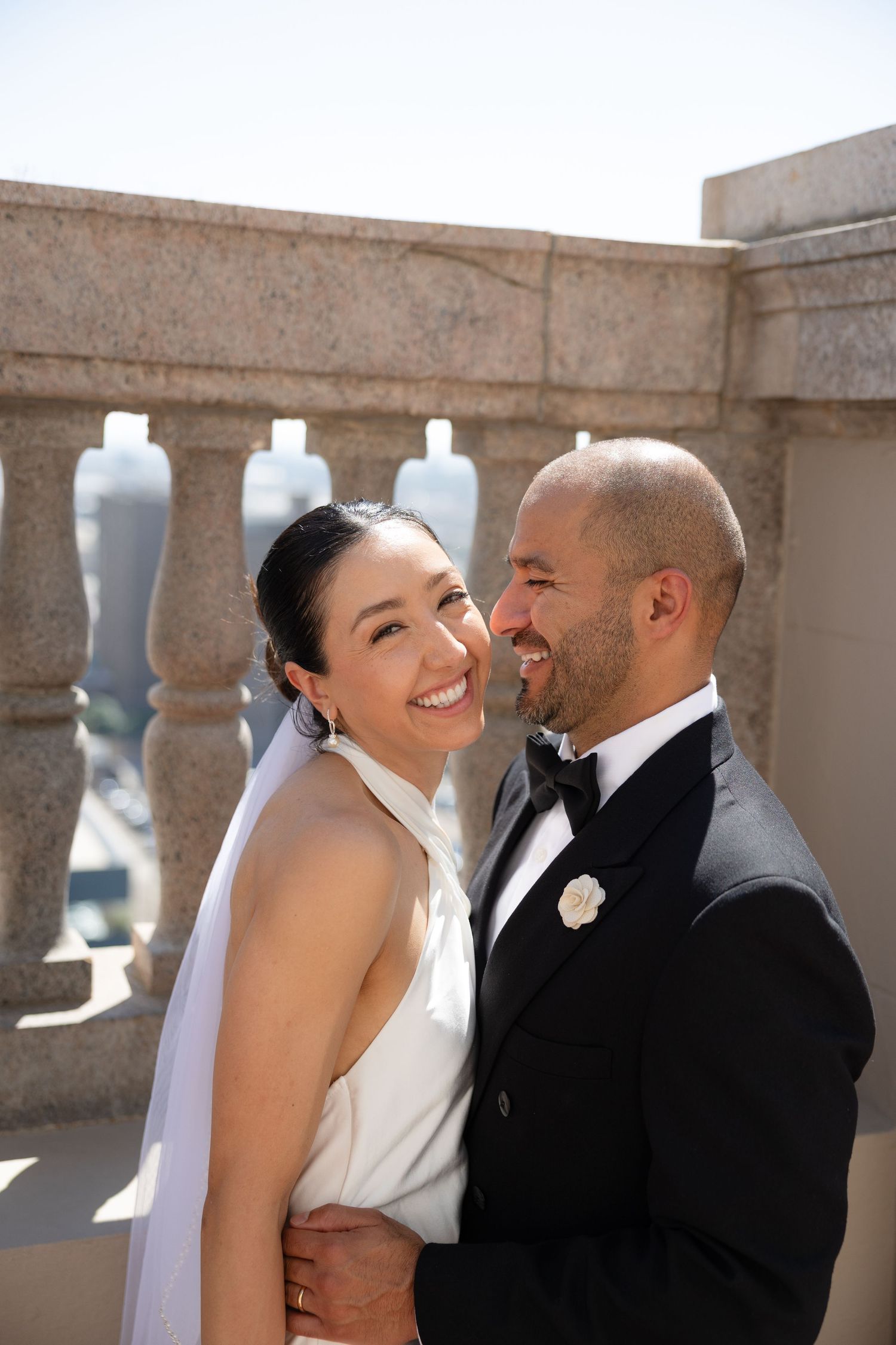Smiling bride and groom embrace on a stone balcony, wearing wedding dress with veil and black tuxedo.