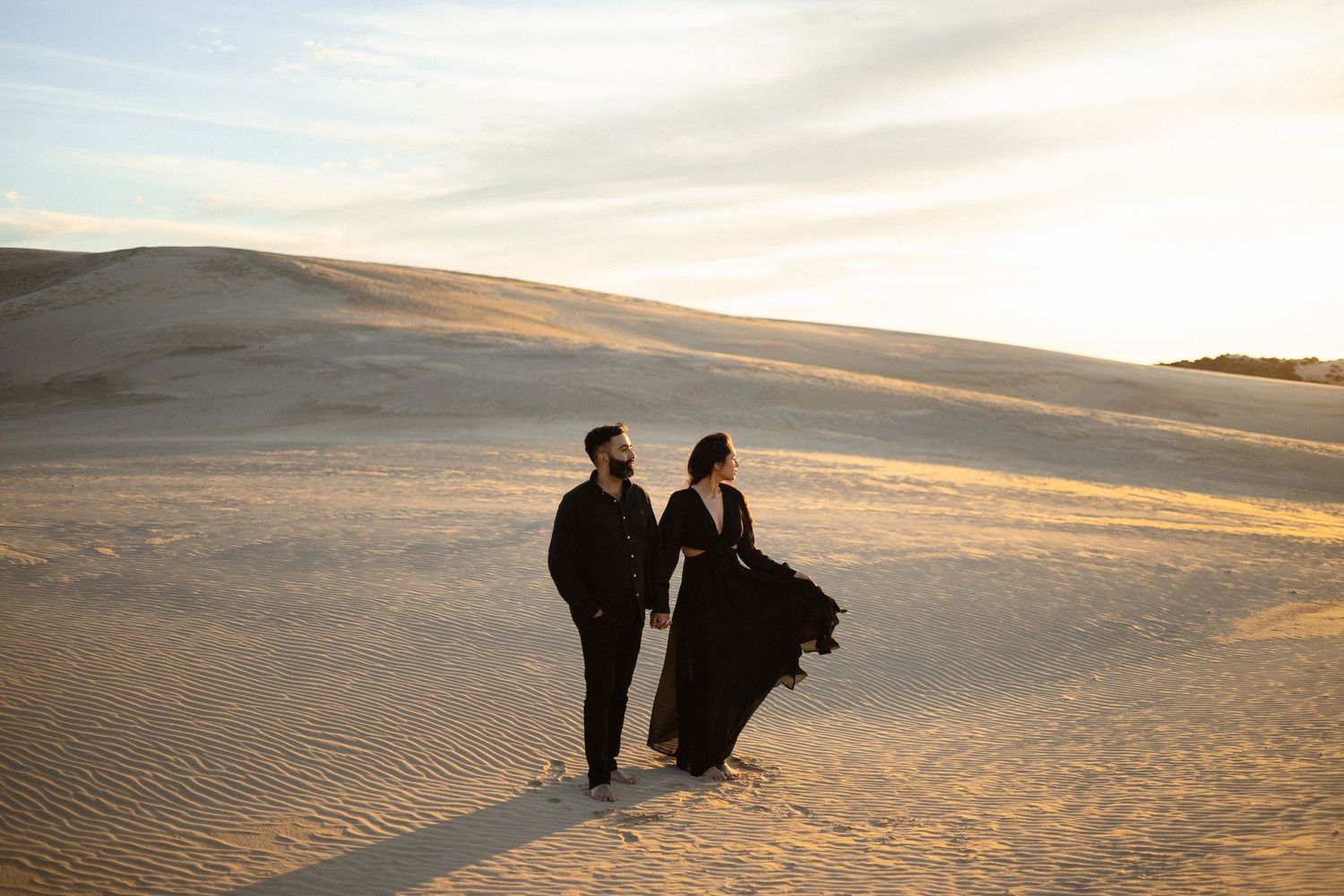 Two silhouetted figures walk together across golden sand dunes at sunset in a desert landscape.