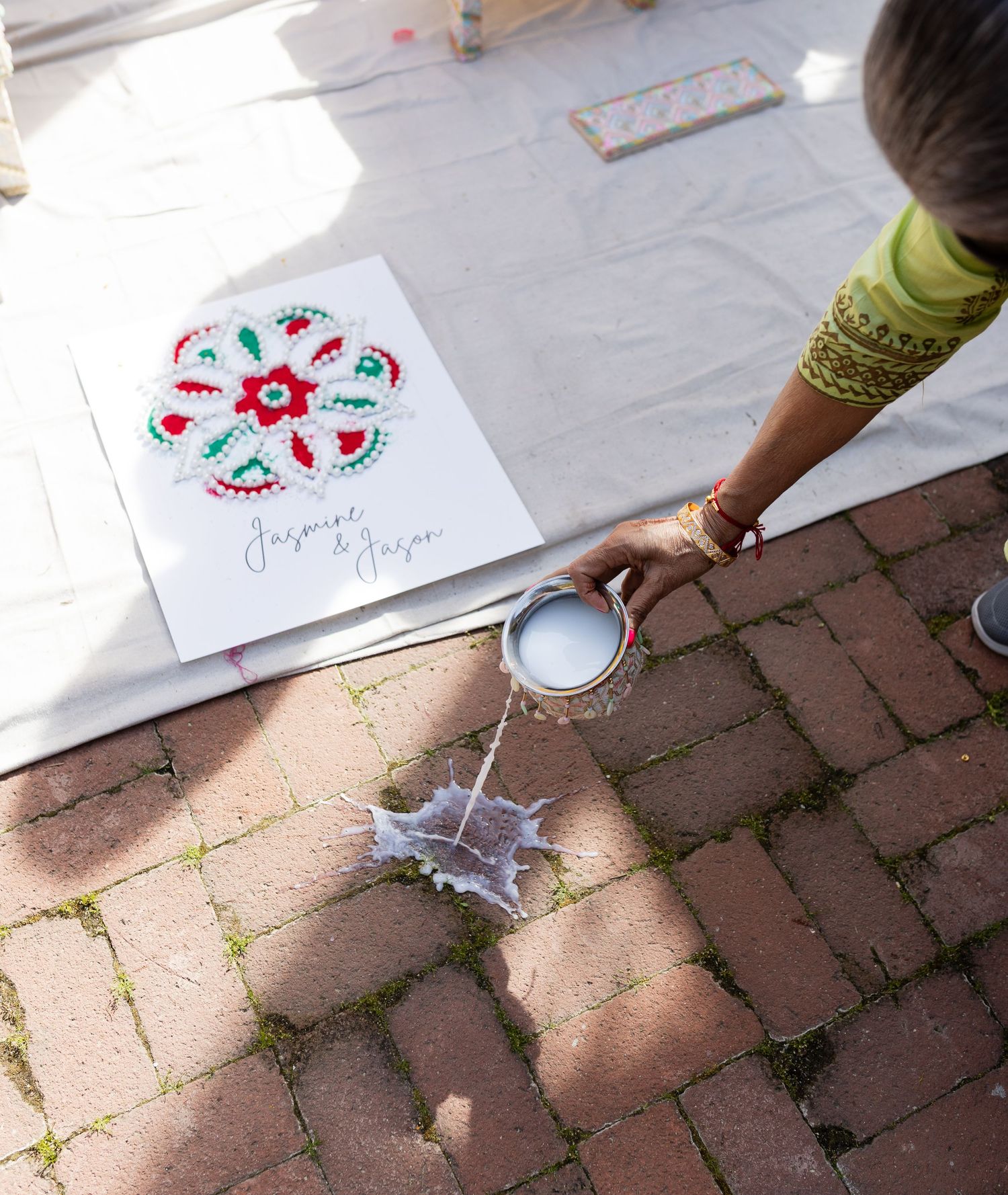 Hands create colorful floral designs on white paper while sitting on brick pavement in sunlight.