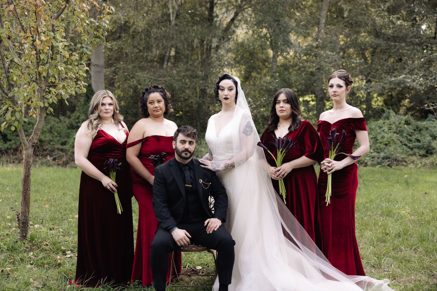Wedding group portrait with burgundy velvet dresses against autumn trees and grass.