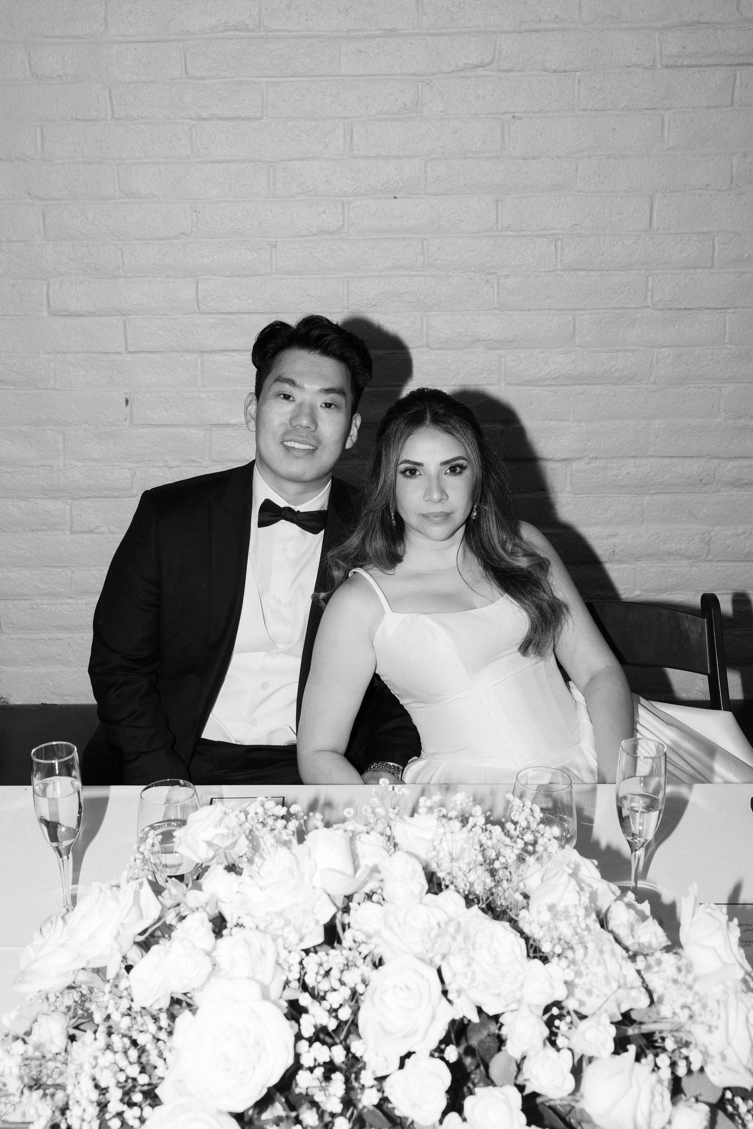 Black and white photo of a couple in wedding attire sitting at a floral reception table.