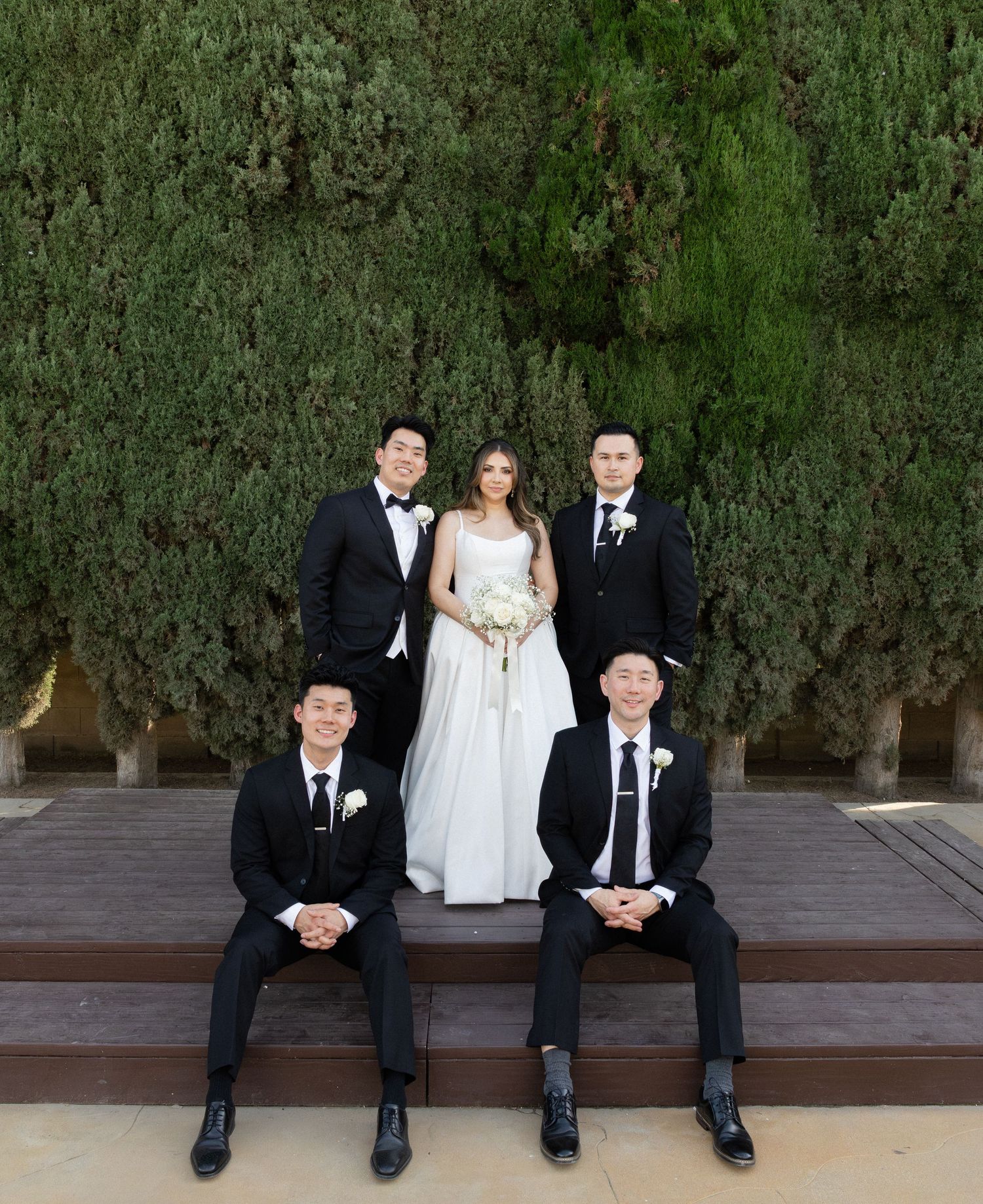 Bride in white gown poses with four groomsmen in black suits before a lush green hedge.